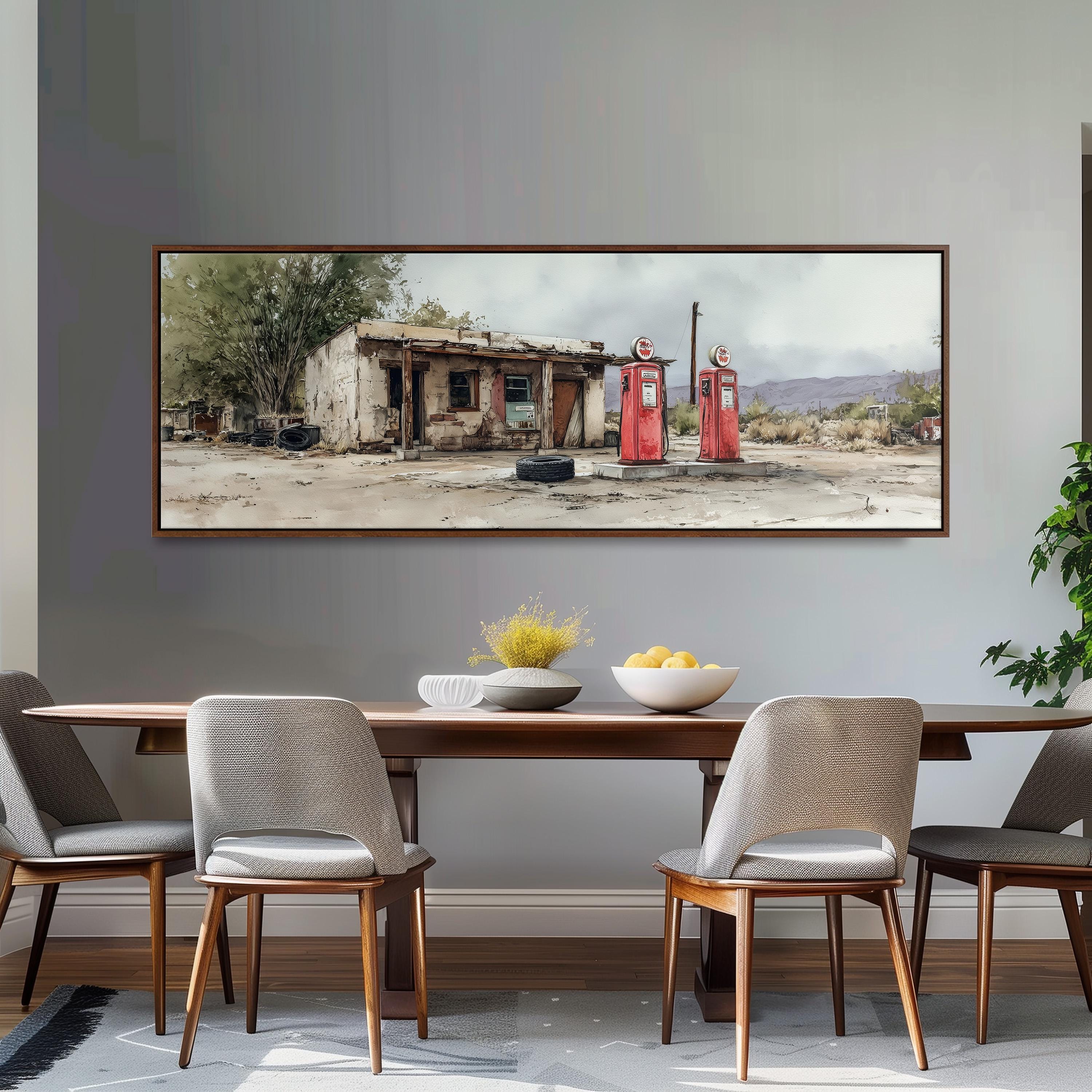 a large painting of a dilapidated building with two red gas pumps in front of it, set against a cloudy sky. The painting is hung on a gray wall above a wooden dining table with four chairs.