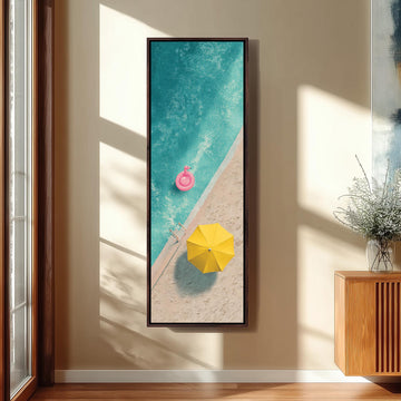 A framed painting depicting a beach scene with a pink inflatable swim toy floating in the turquoise water, a yellow umbrella, and a sandy beach.