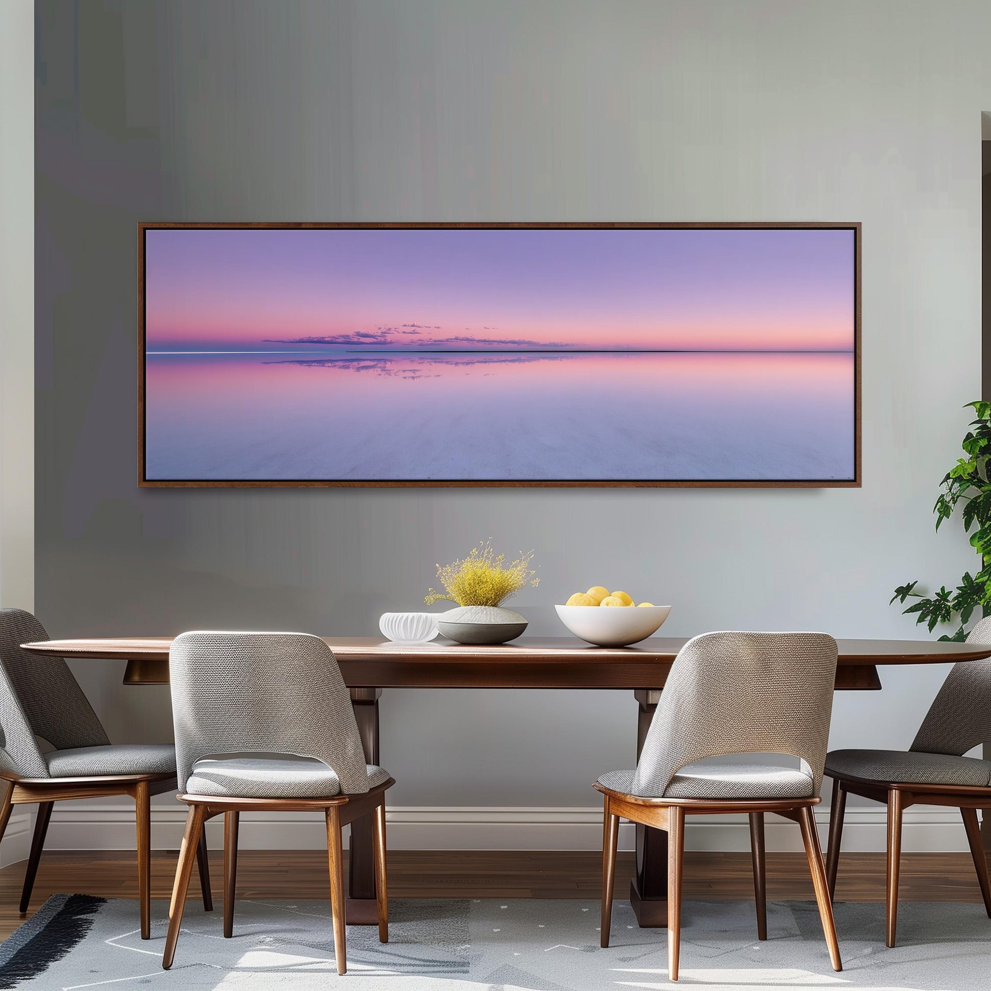 a dining room with a large, rectangular painting of a sunset over a calm body of water hanging on the wall above a wooden dining table. The table is set with a bowl of fruit and a vase of flowers, creating a cozy and inviting atmosphere.