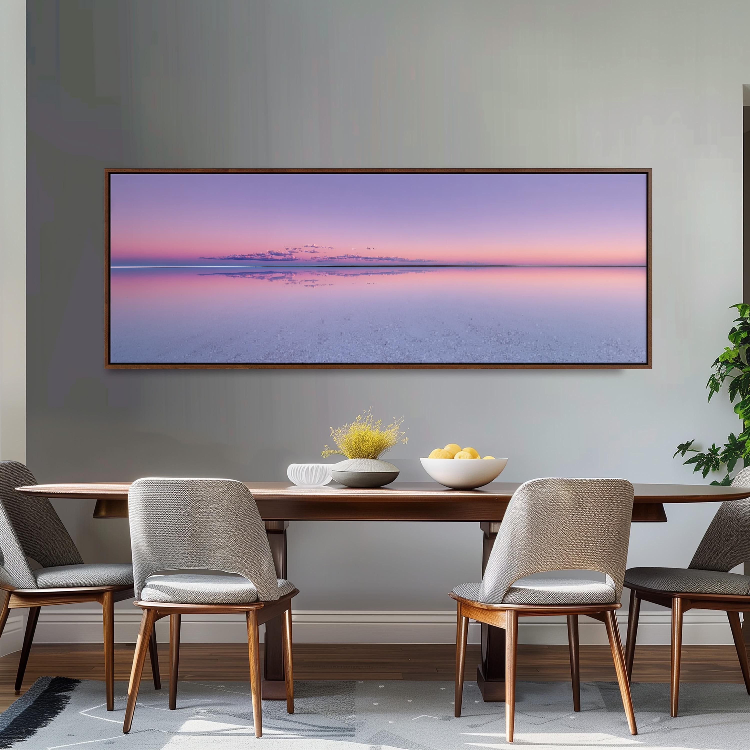 a dining room with a large, rectangular painting of a sunset over a calm body of water hanging on the wall above a wooden dining table. The table is set with a bowl of fruit and a vase of flowers, creating a cozy and inviting atmosphere.