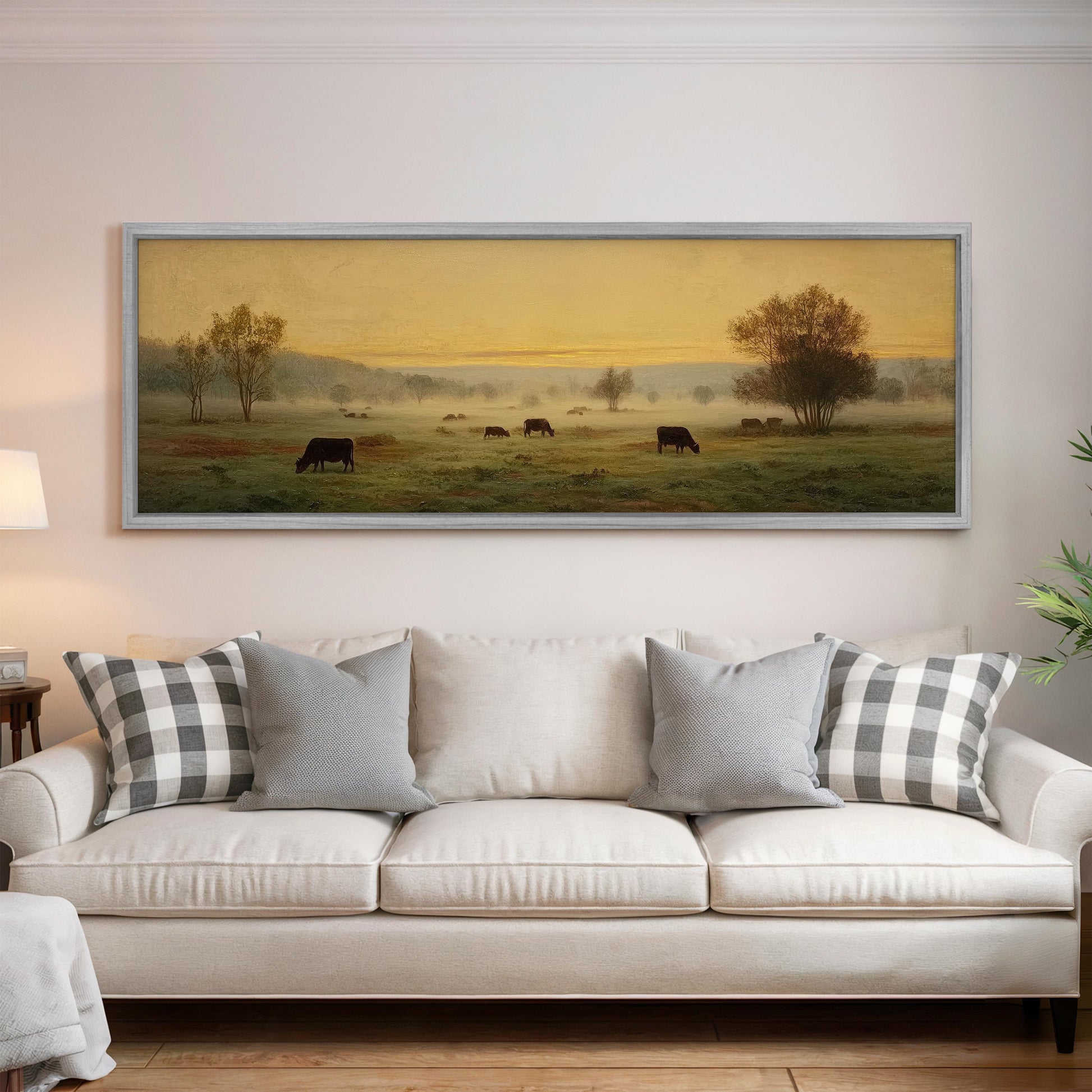 a large painting of a pastoral scene with cows grazing in a field, hanging on a wall above a white sofa with patterned throw pillows.