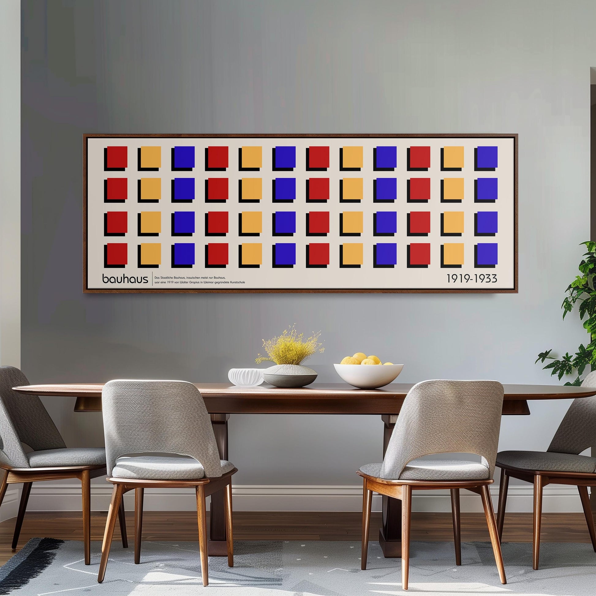 a dining room with a wooden table and four chairs. Above the table, there is a large framed poster with a repeating pattern of squares in various colors. The poster is hanging on a gray wall, and the dining table is set with a bowl of fruit and a vase.