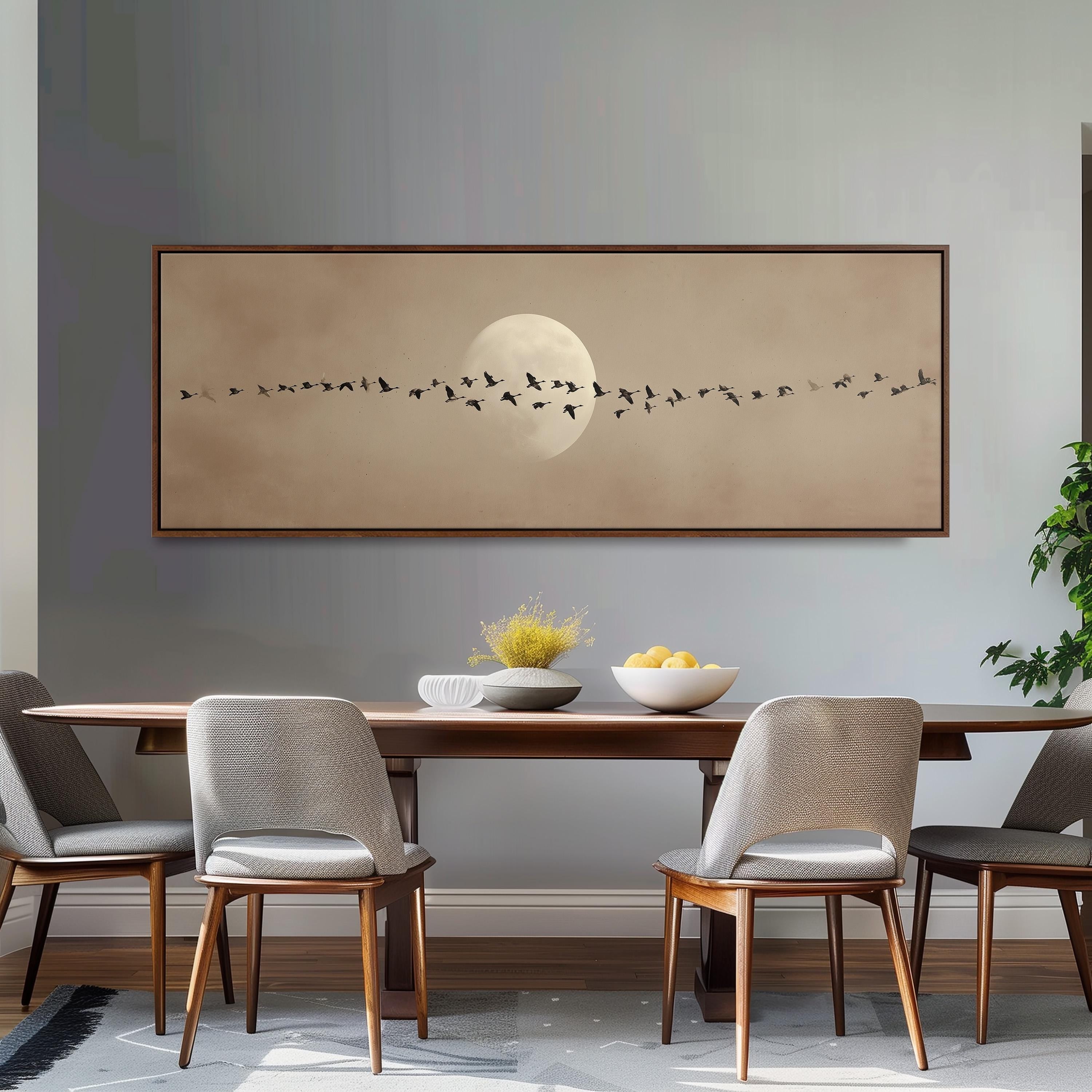 a dining room with a wooden table and four chairs, a large framed artwork depicting a flock of birds flying over a moonlit sky, and a bowl of fruit on the table.