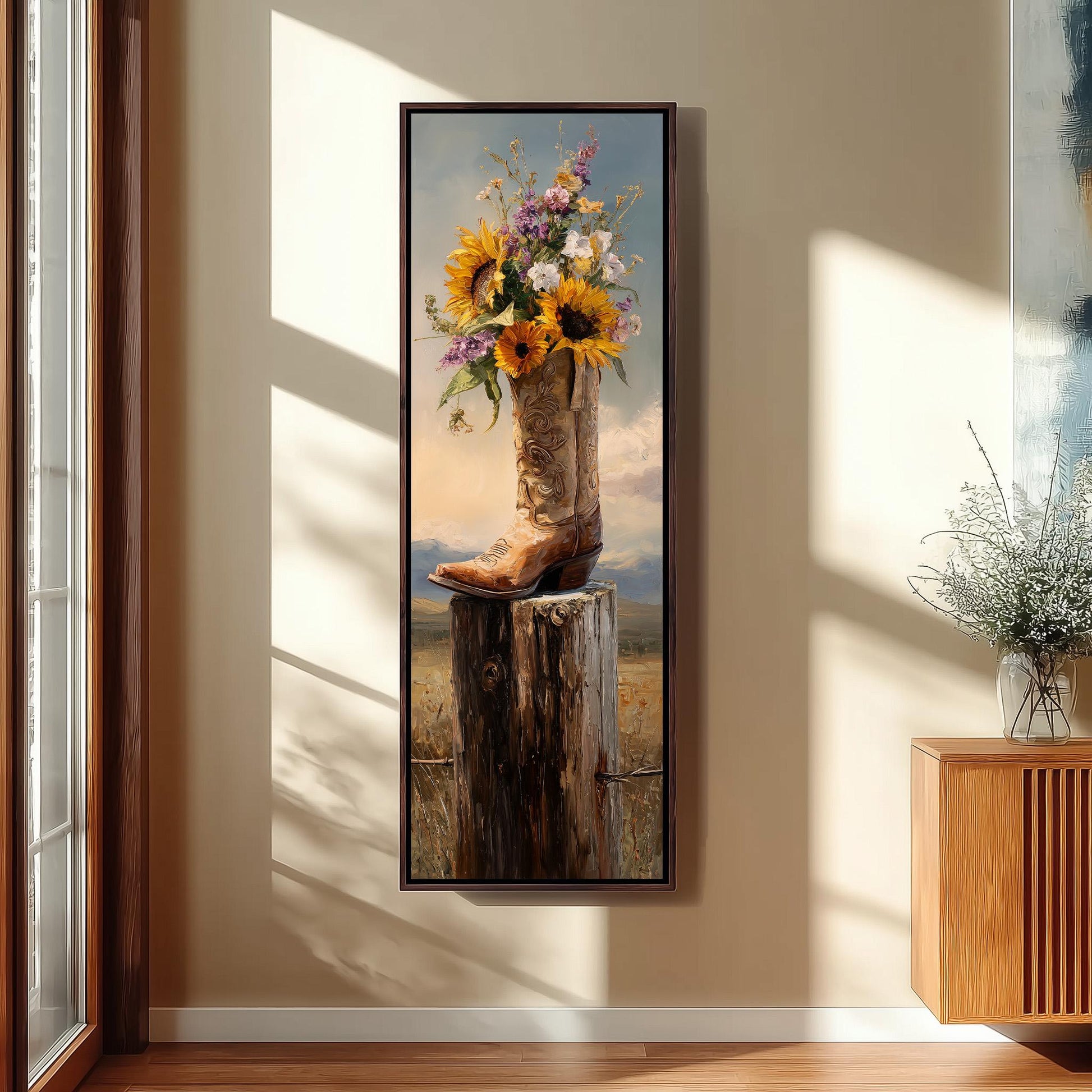 A framed painting of a cowboy boot filled with flowers is hanging on a wall in a room with a wooden floor and a window.