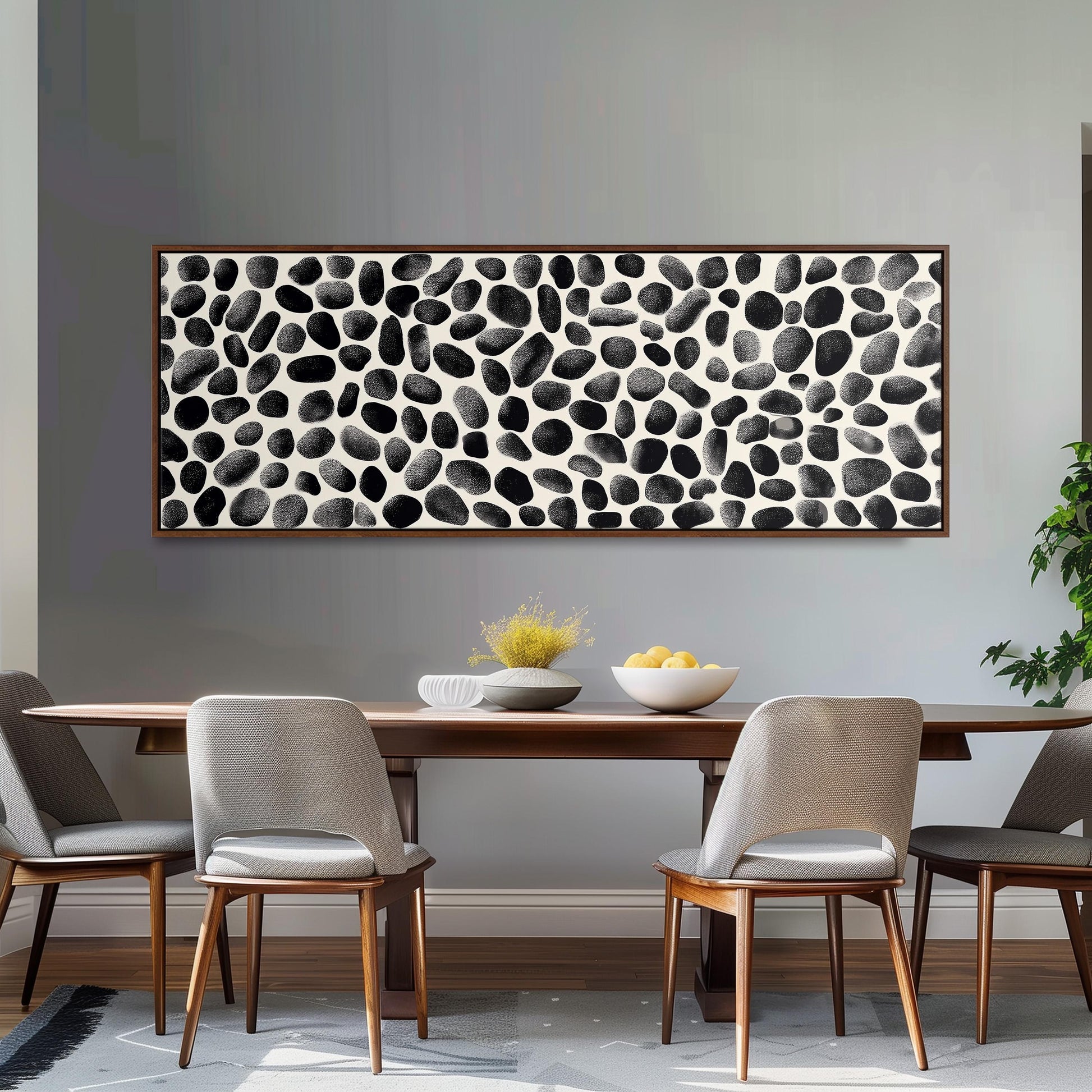 a dining room with a wooden table and four chairs. Above the table, there is a large black and white abstract painting with a repeating pattern of black and white circles. The table is set with a bowl of fruit and a vase of flowers, creating a cozy and inviting atmosphere.