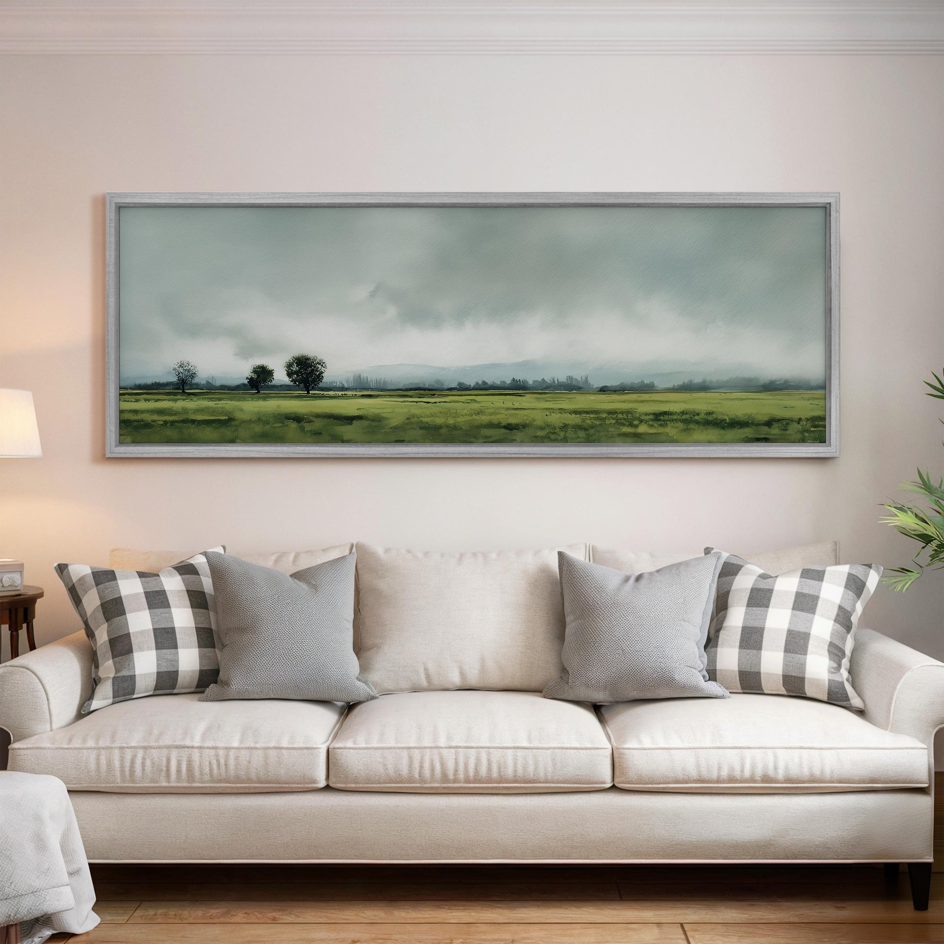 a living room with a white sofa adorned with gray and white patterned pillows. Above the sofa, a large landscape painting depicting a countryside scene with trees and rolling hills is displayed on the wall.