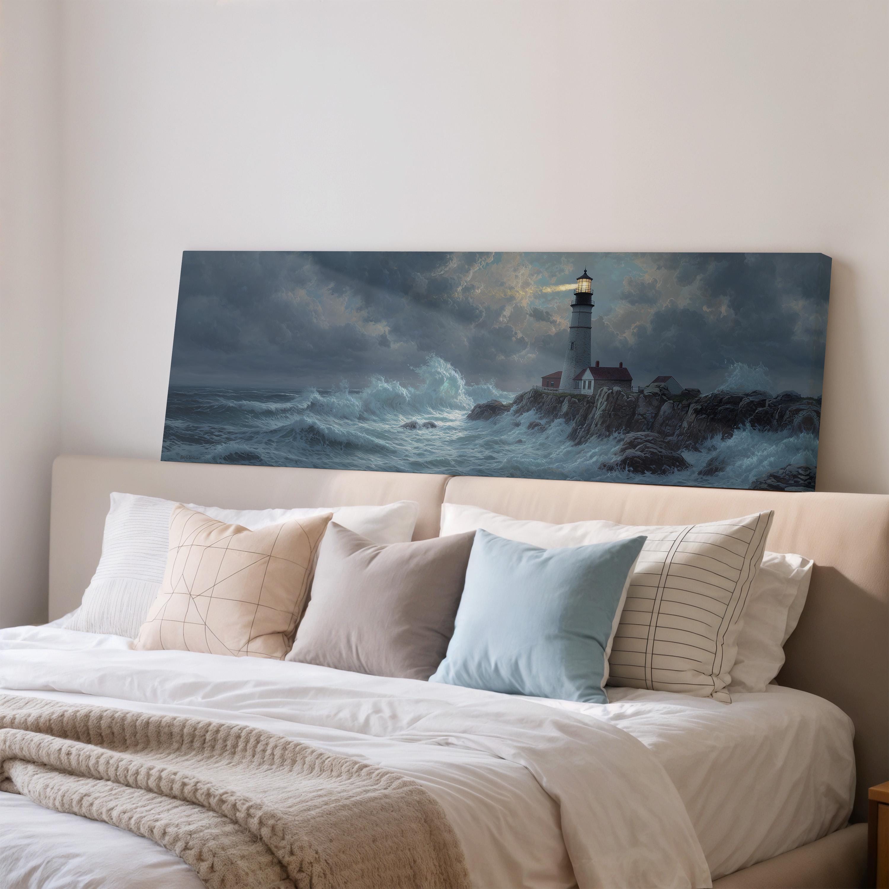a large painting of a lighthouse on a rocky shore, hanging on a wall above a bed with white pillows and a gray blanket.