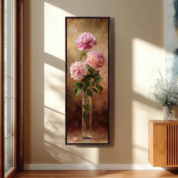 A framed painting of three pink peonies in a vase is hanging on a wall in a room with a wooden cabinet and a window.