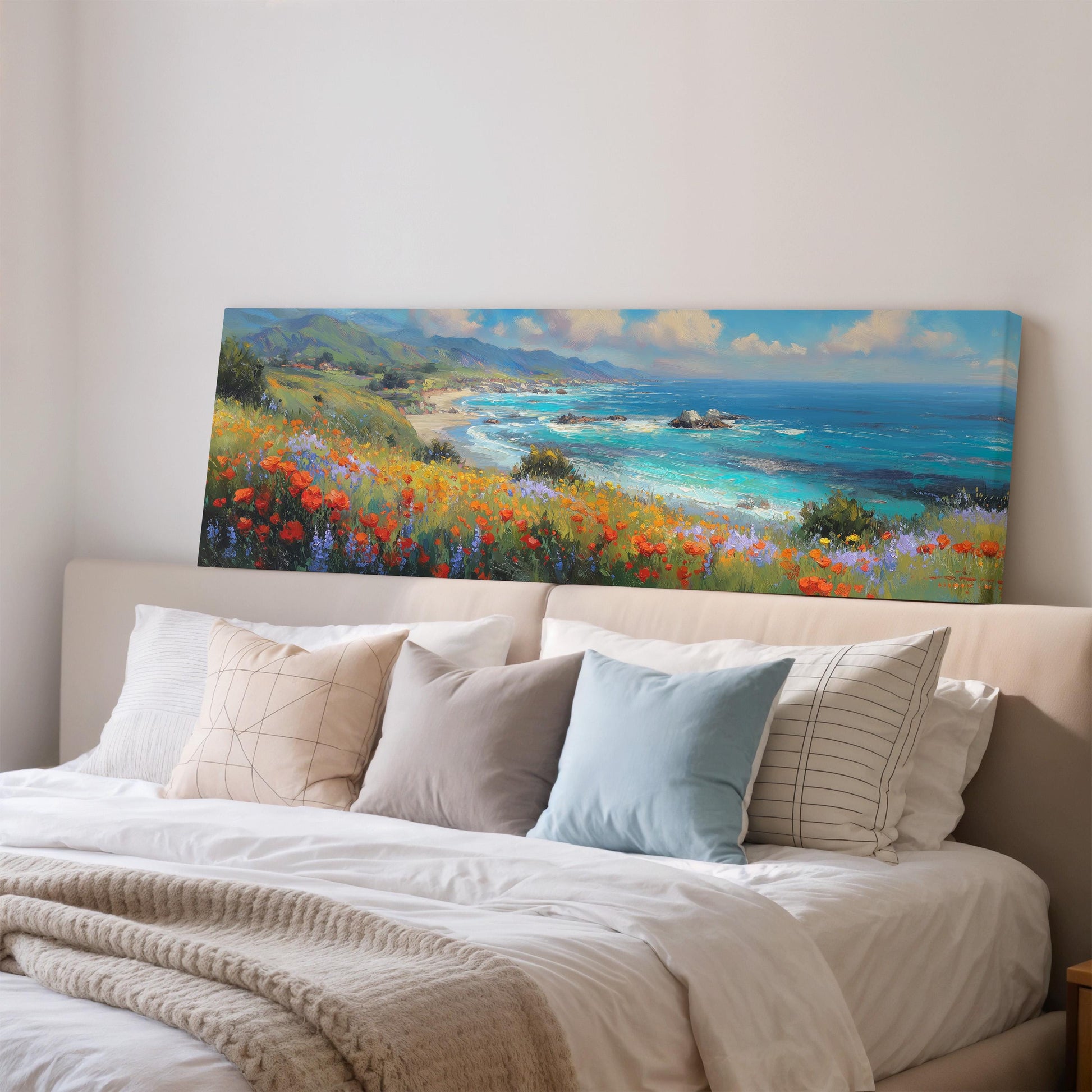 a large painting of a coastal landscape with a beach, mountains, and a blue ocean, hanging on a wall above a bed with white pillows and a gray blanket.