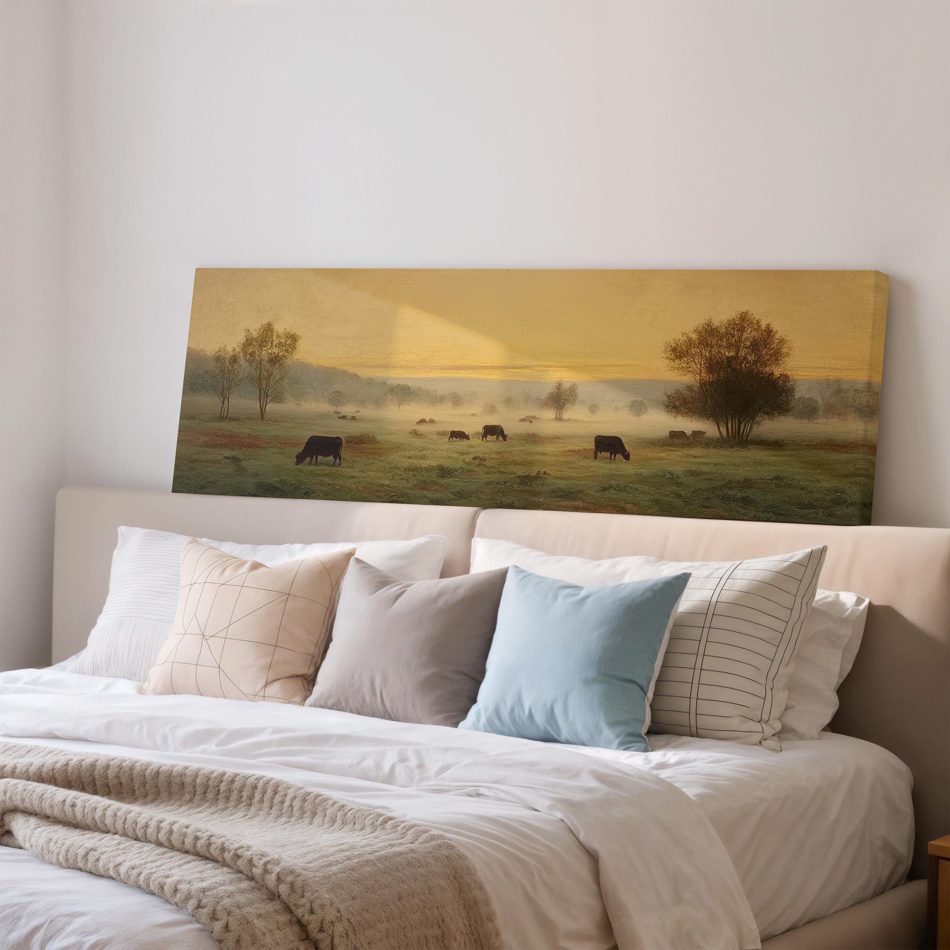 a large painting of a pastoral scene with cows grazing in a field, hanging on a wall above a bed with white pillows and a gray blanket.
