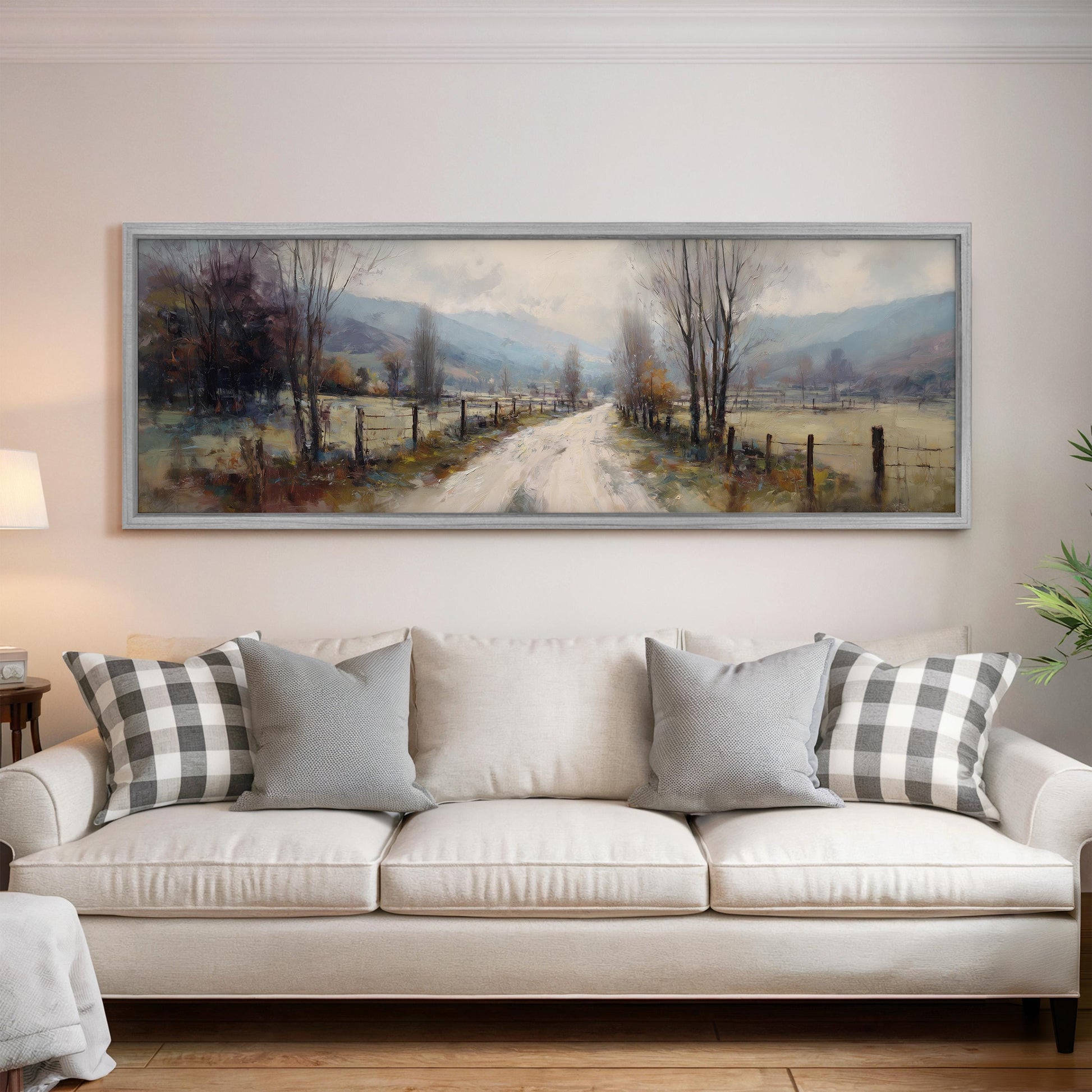 a large painting of a country road with trees and a fence, hanging on a wall above a white sofa with patterned throw pillows.