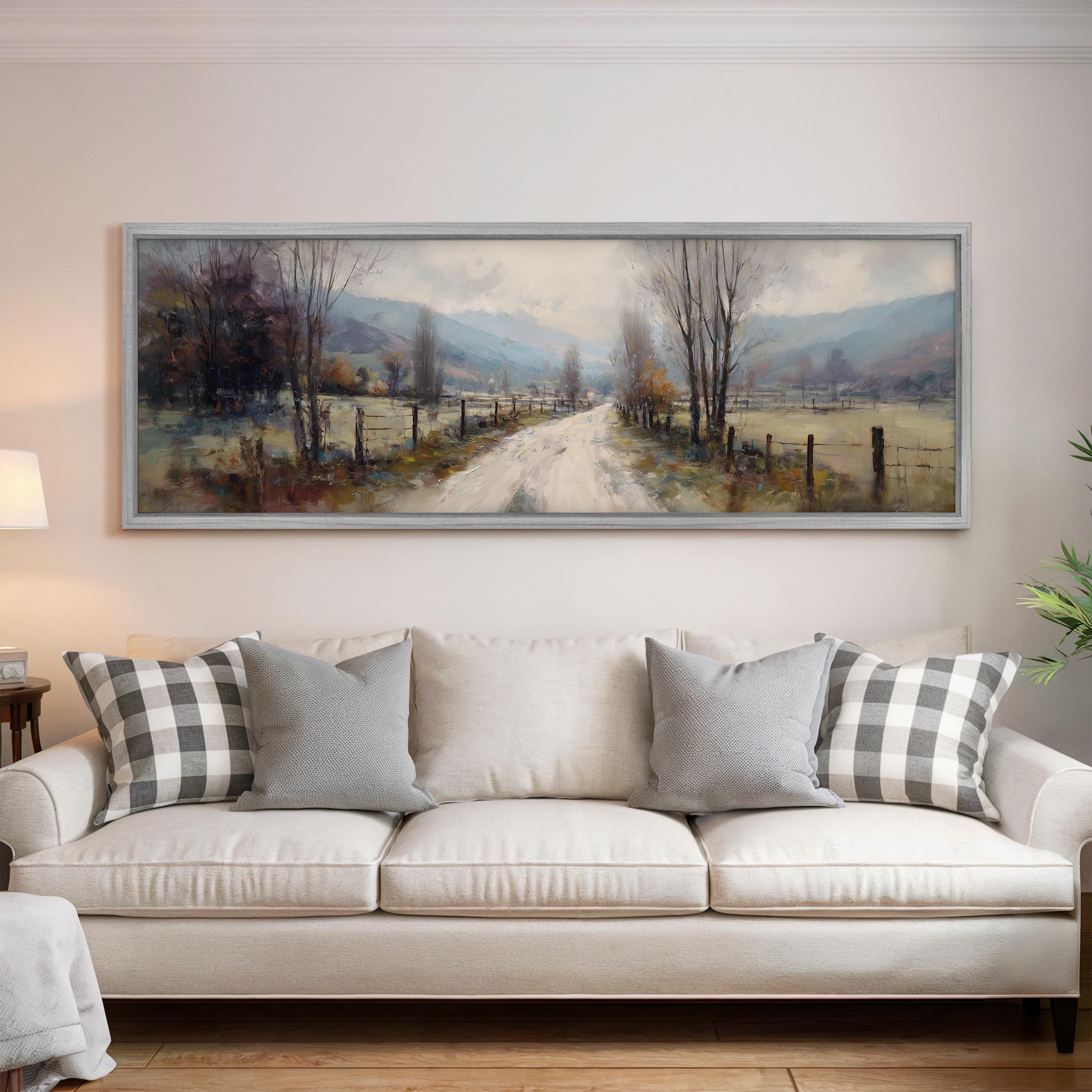 a large painting of a country road with trees and a fence, hanging on a wall above a white sofa with patterned throw pillows.
