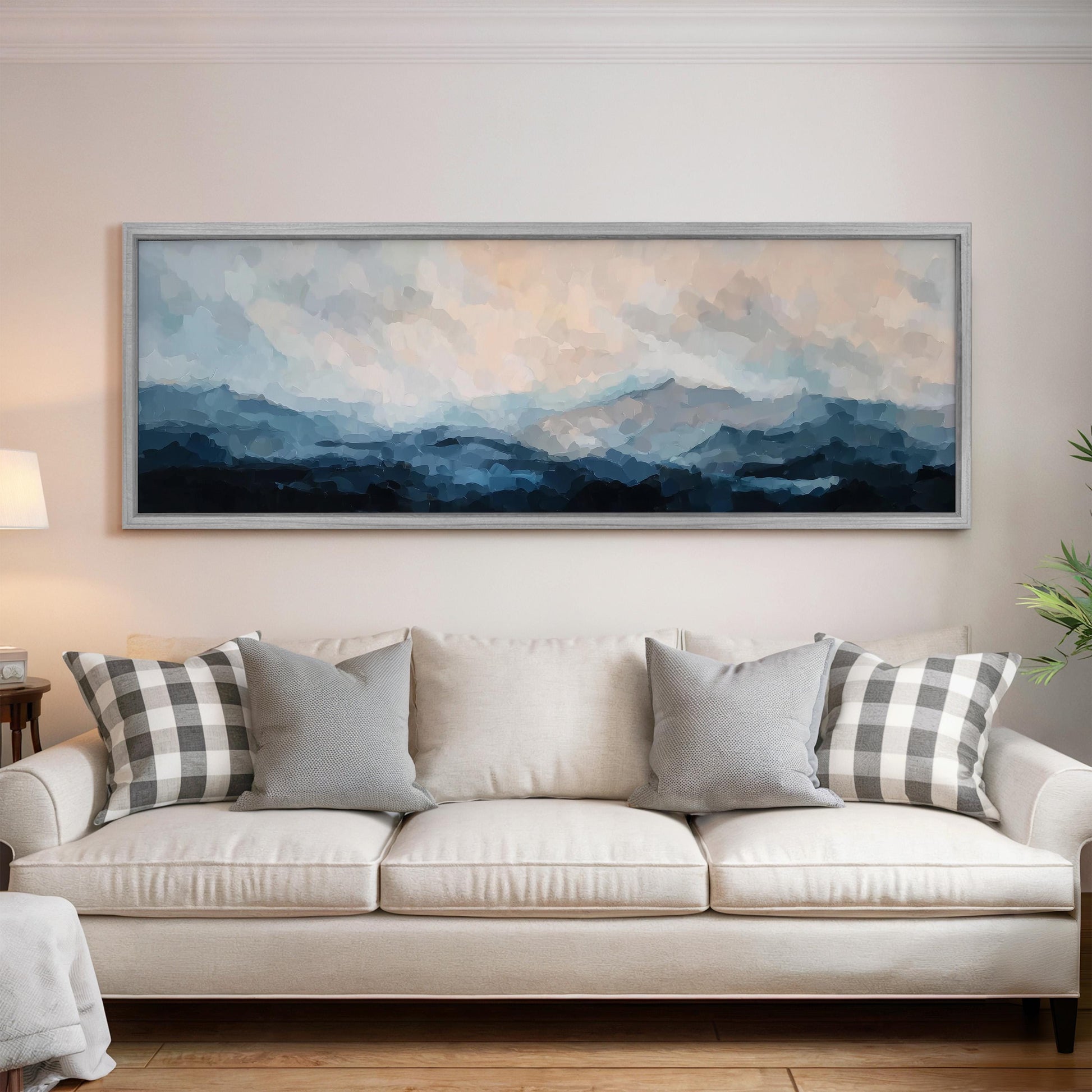 a large, framed painting of a mountainous landscape hanging on a wall above a white sofa with gray and white patterned throw pillows.