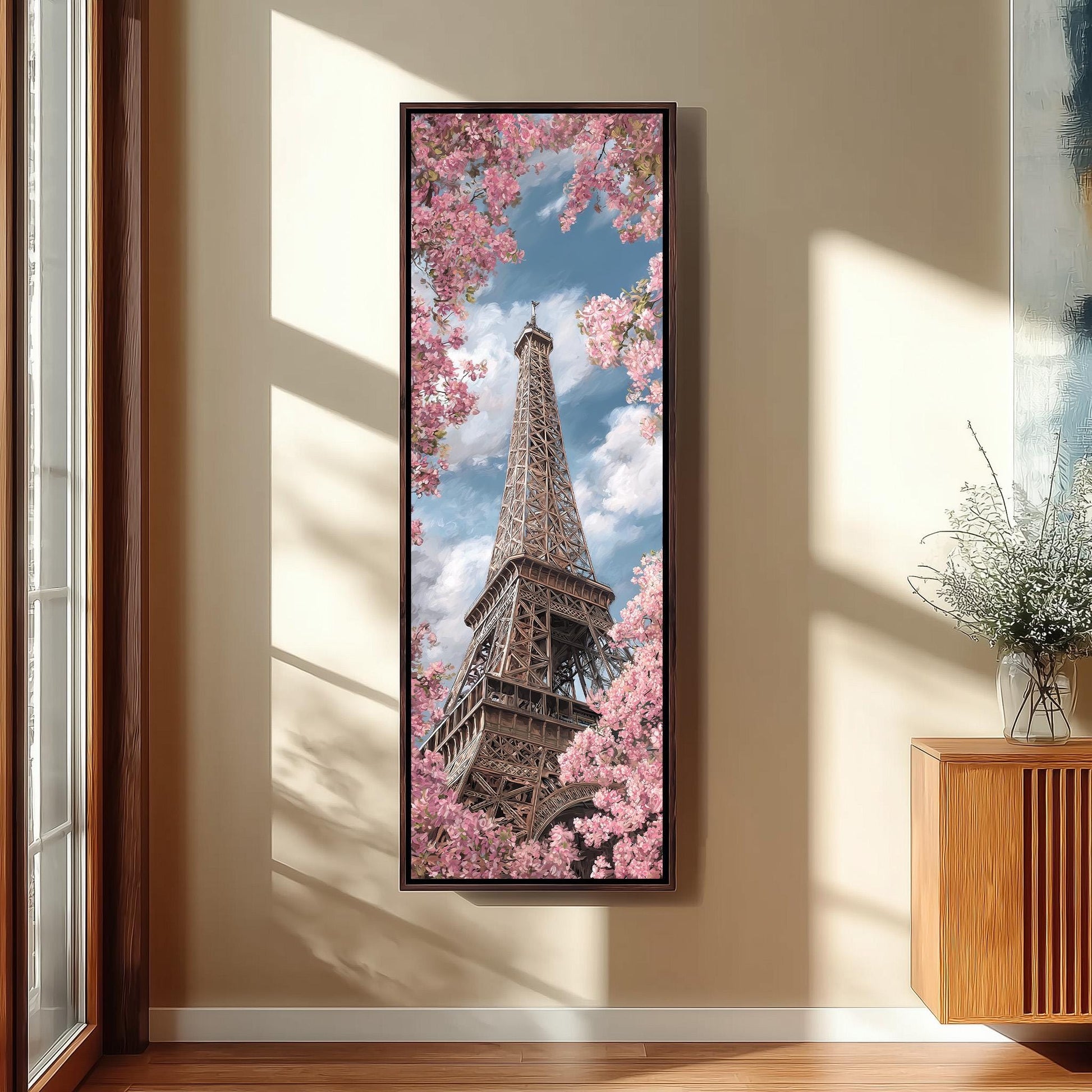 A framed photograph of the Eiffel Tower in Paris, France, surrounded by pink cherry blossoms, is displayed on the wall in a room with a wooden cabinet and a vase of flowers.
