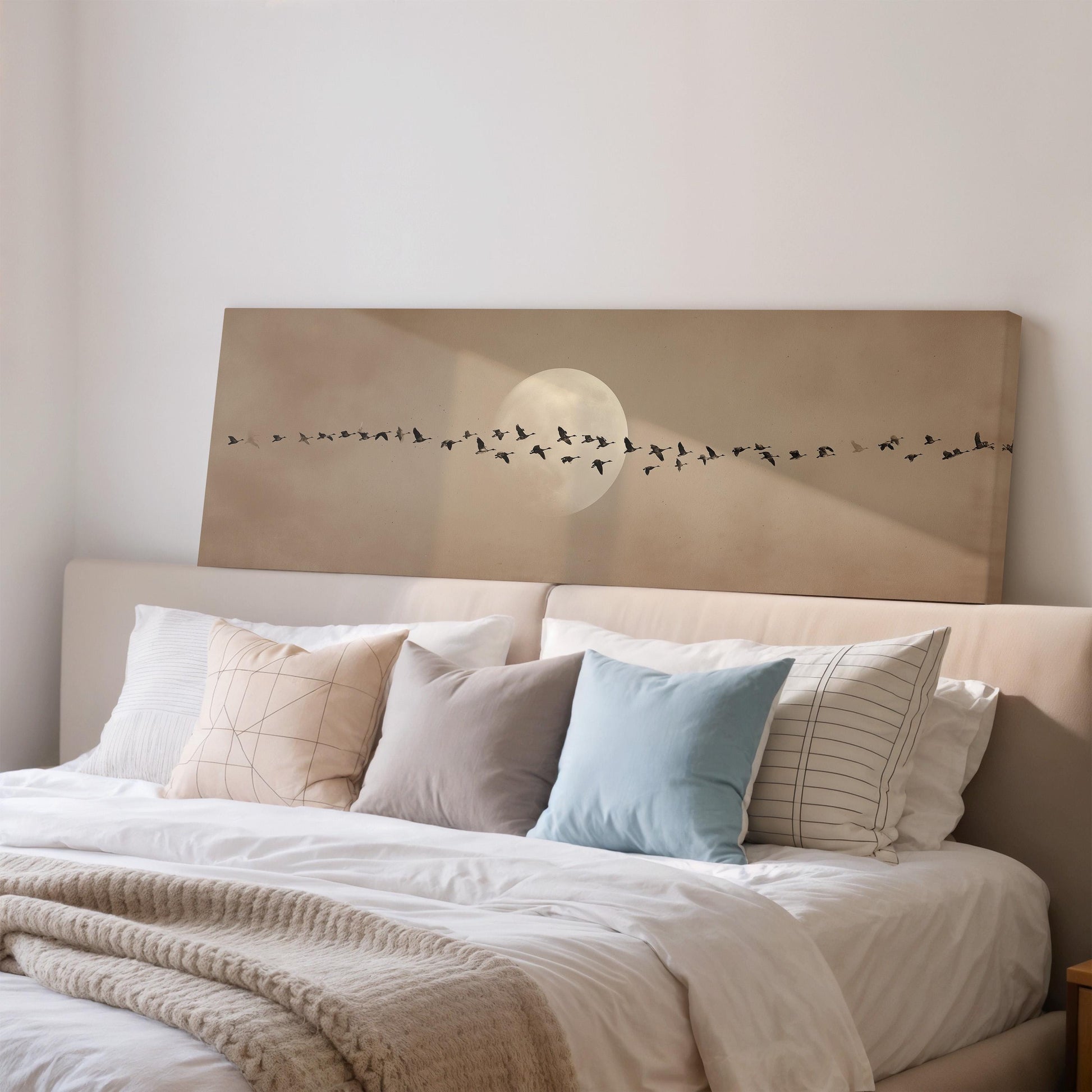 a cozy bedroom with a large bed adorned with white sheets and a blue pillow. Above the bed, there is a framed artwork depicting a flock of birds flying in formation, with a large moon in the background.