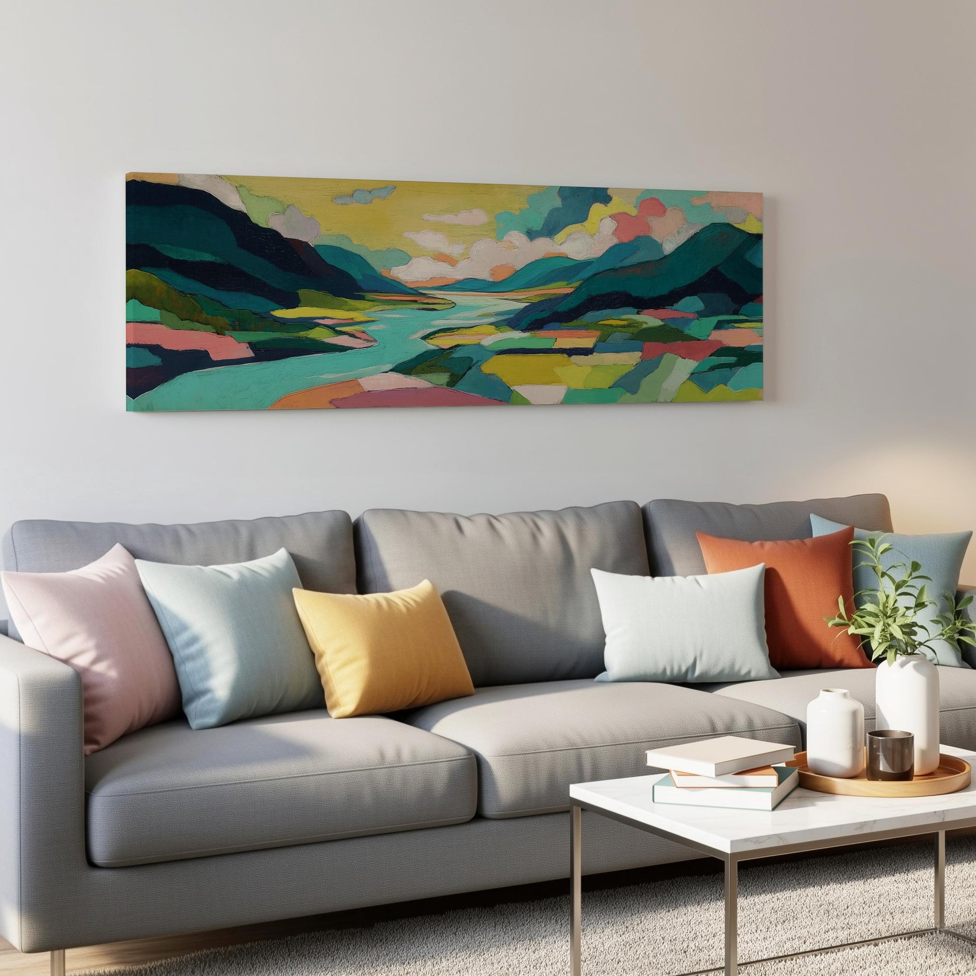 a modern living room with a gray sofa adorned with colorful throw pillows. Above the sofa, a large landscape painting depicting a serene mountain scene is displayed on the wall. The room also includes a coffee table with books and a plant, creating a cozy and inviting atmosphere.