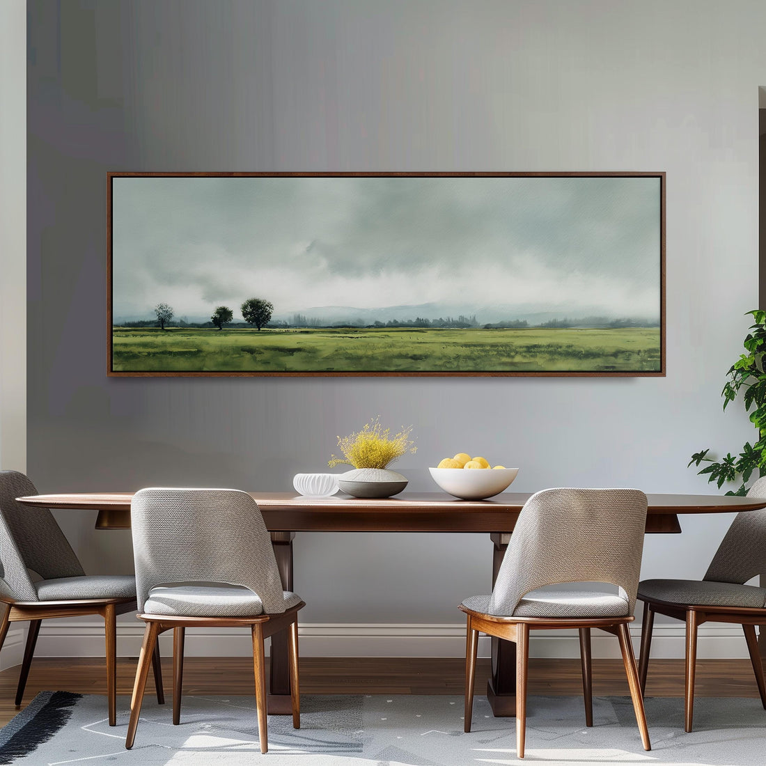 a dining room with a large painting of a field hanging on the wall above a wooden dining table with four chairs. The painting depicts a serene landscape with a cloudy sky and rolling hills.