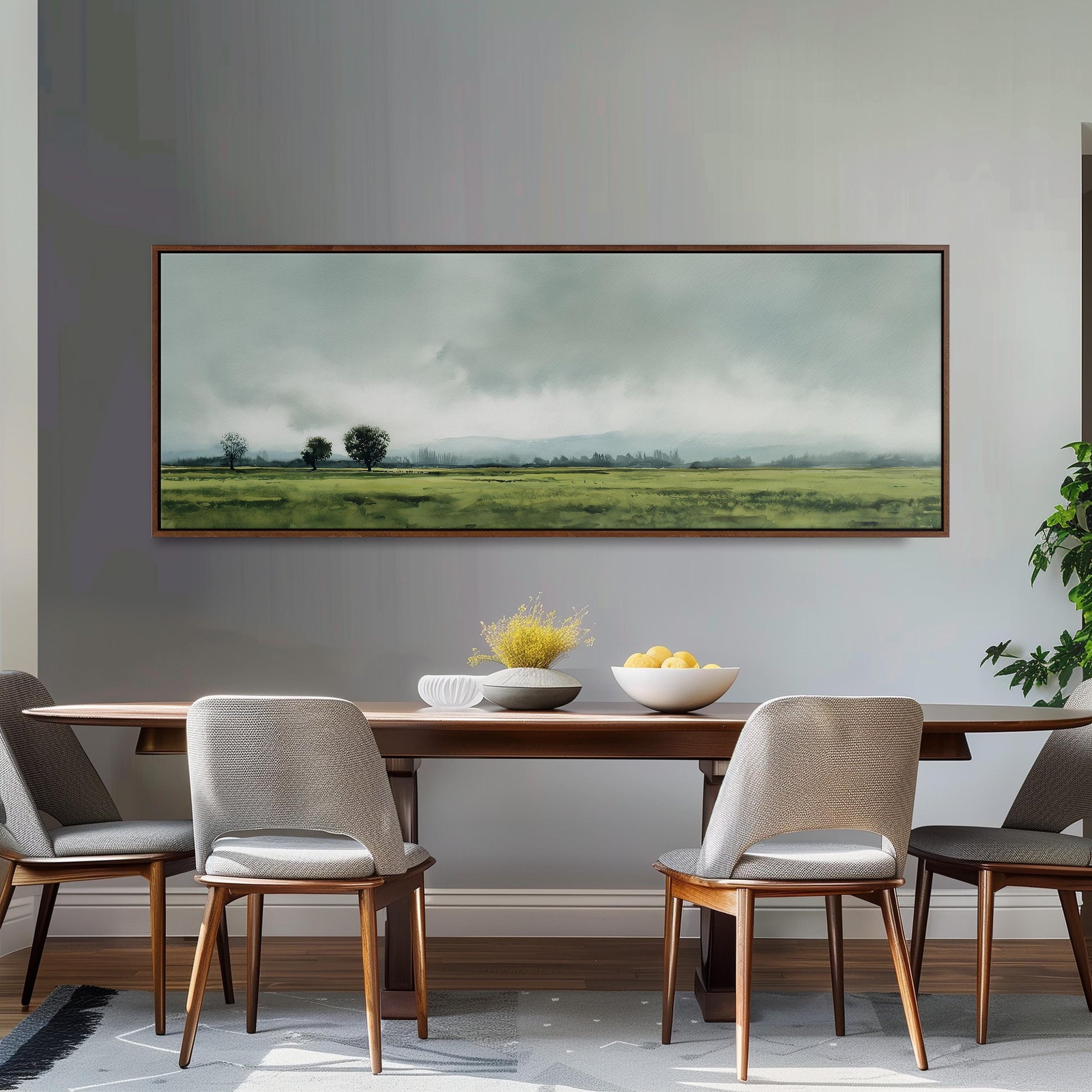 a dining room with a large painting of a field hanging on the wall above a wooden dining table with four chairs. The painting depicts a serene landscape with a cloudy sky and rolling hills.