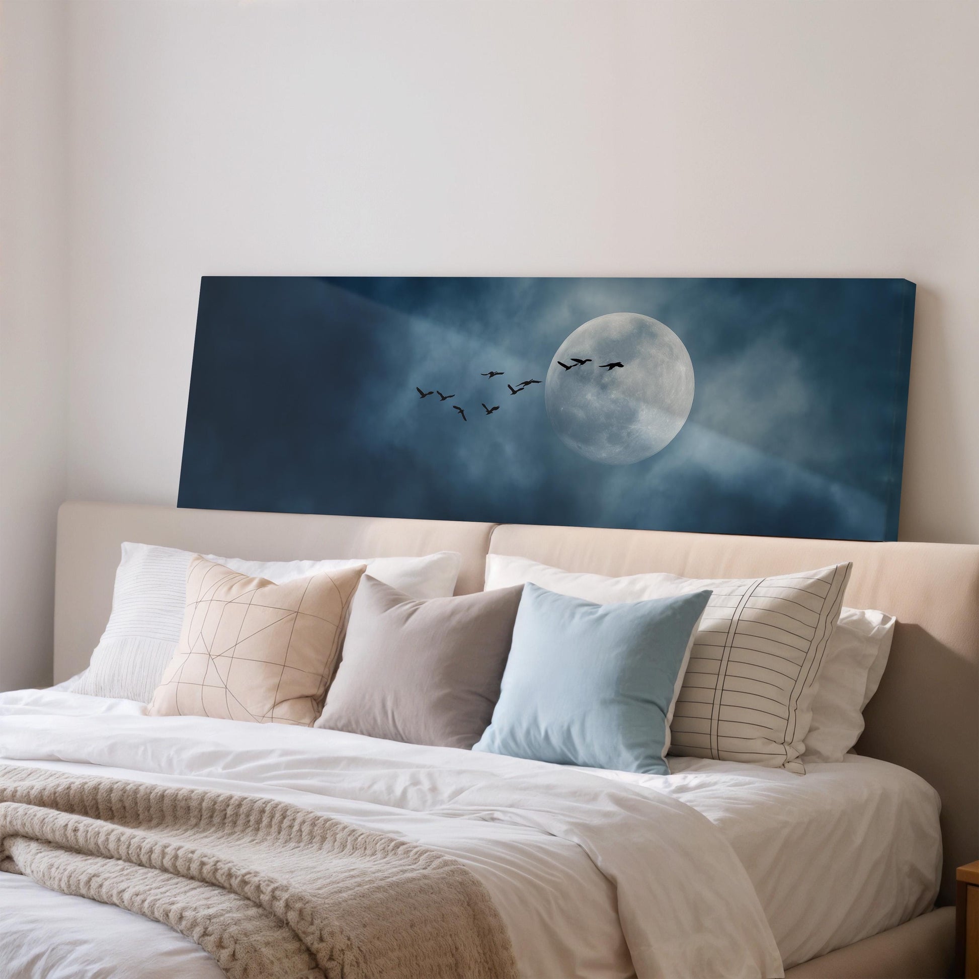 a serene bedroom scene with a large bed adorned with white linens and a blue comforter. Above the bed, a large canvas print features a night sky with a full moon and a flock of birds flying across it.