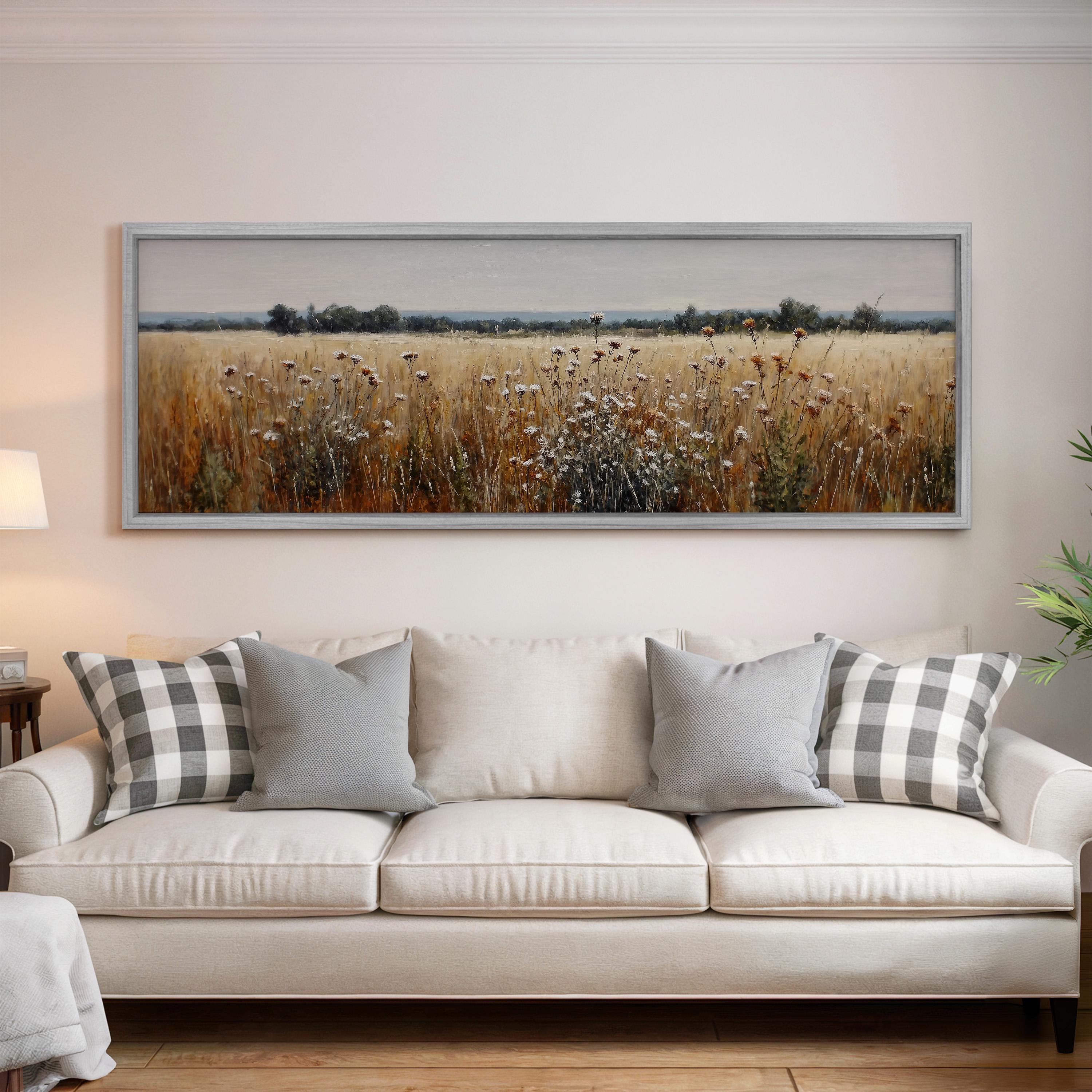 a large, framed painting of a field with tall grass and wildflowers hanging on a wall above a white sofa with gray and white patterned pillows.