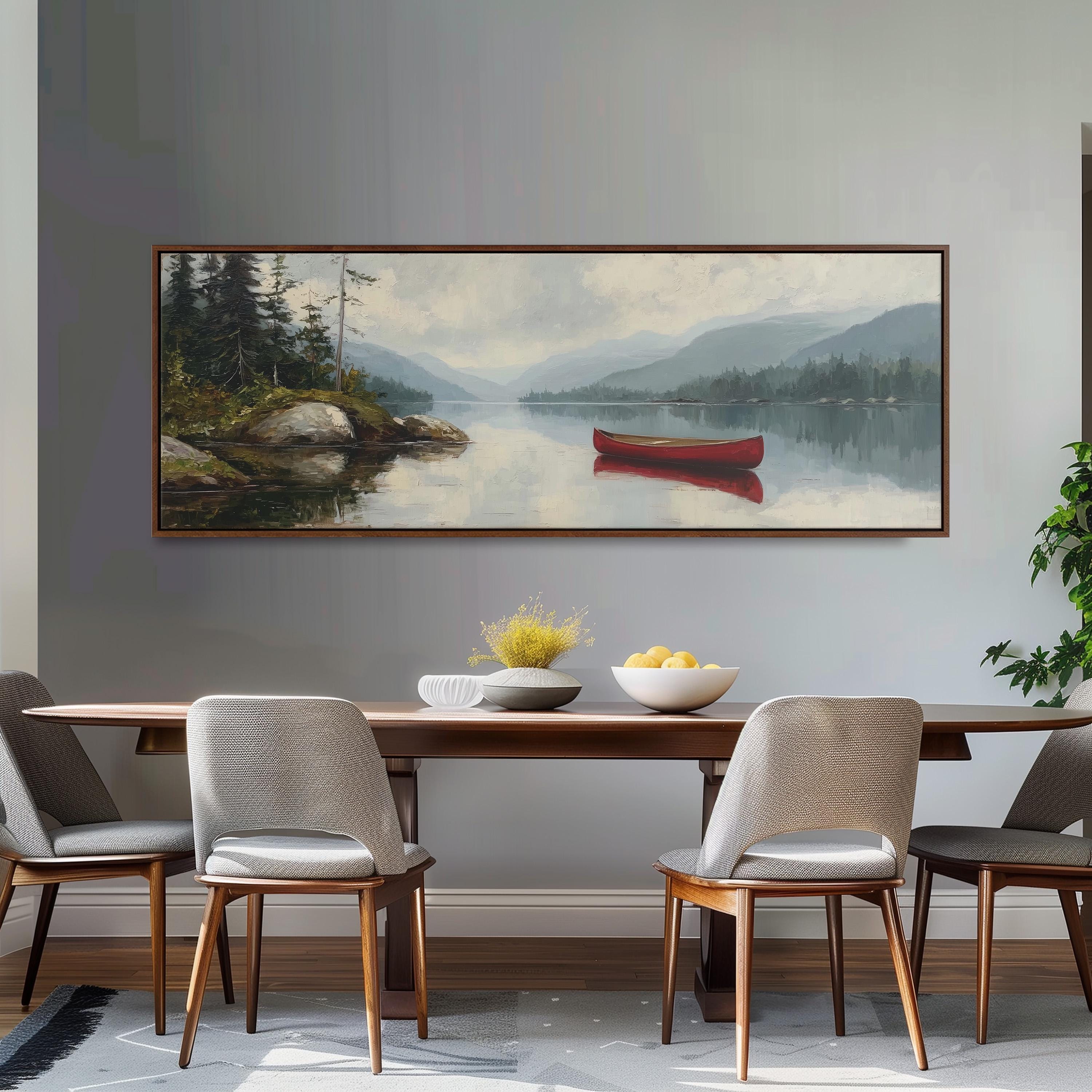 a large painting of a serene lake scene with a red canoe on the water, surrounded by mountains and trees. The painting is hung on a gray wall above a wooden dining table with four chairs. The table is set with a bowl of fruit and a vase of flowers, creating a cozy and inviting atmosphere.