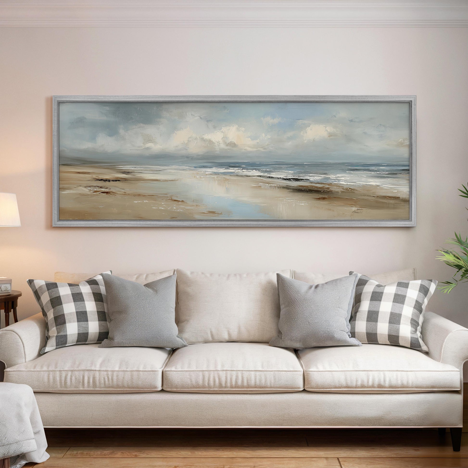 a large, framed painting of a beach scene hanging on a wall above a white sofa with gray and white patterned throw pillows.