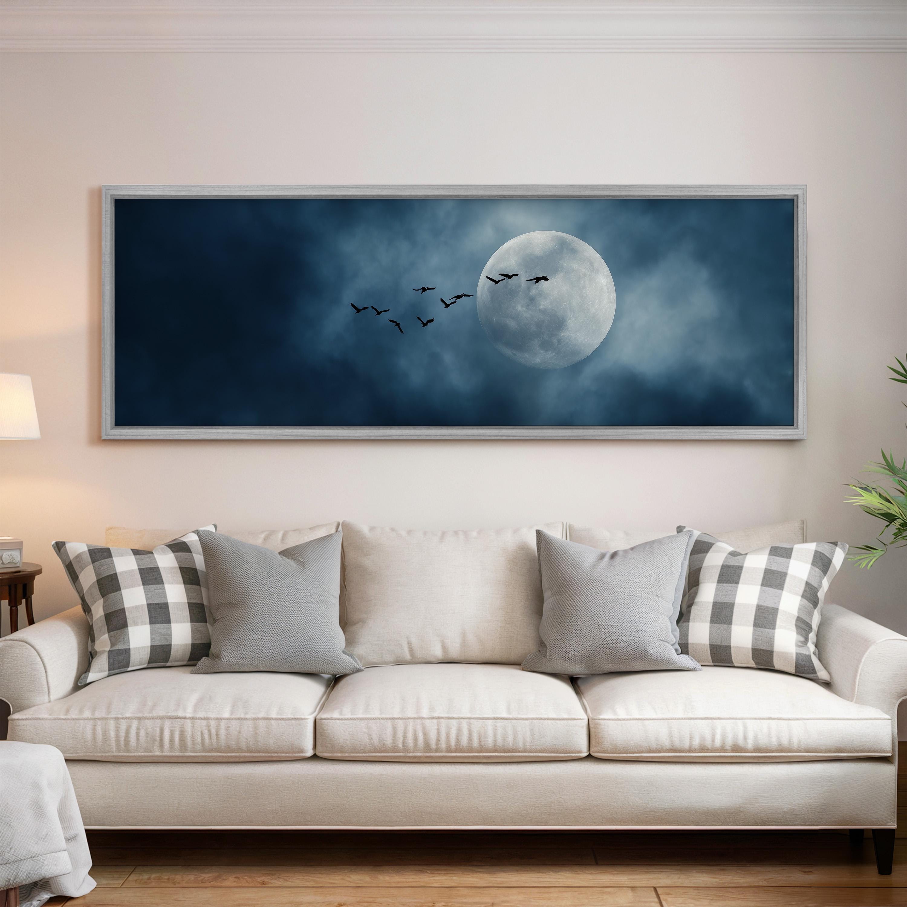 a large framed picture of a full moon and a flock of birds flying in the night sky, hanging on a wall above a white couch with patterned throw pillows.