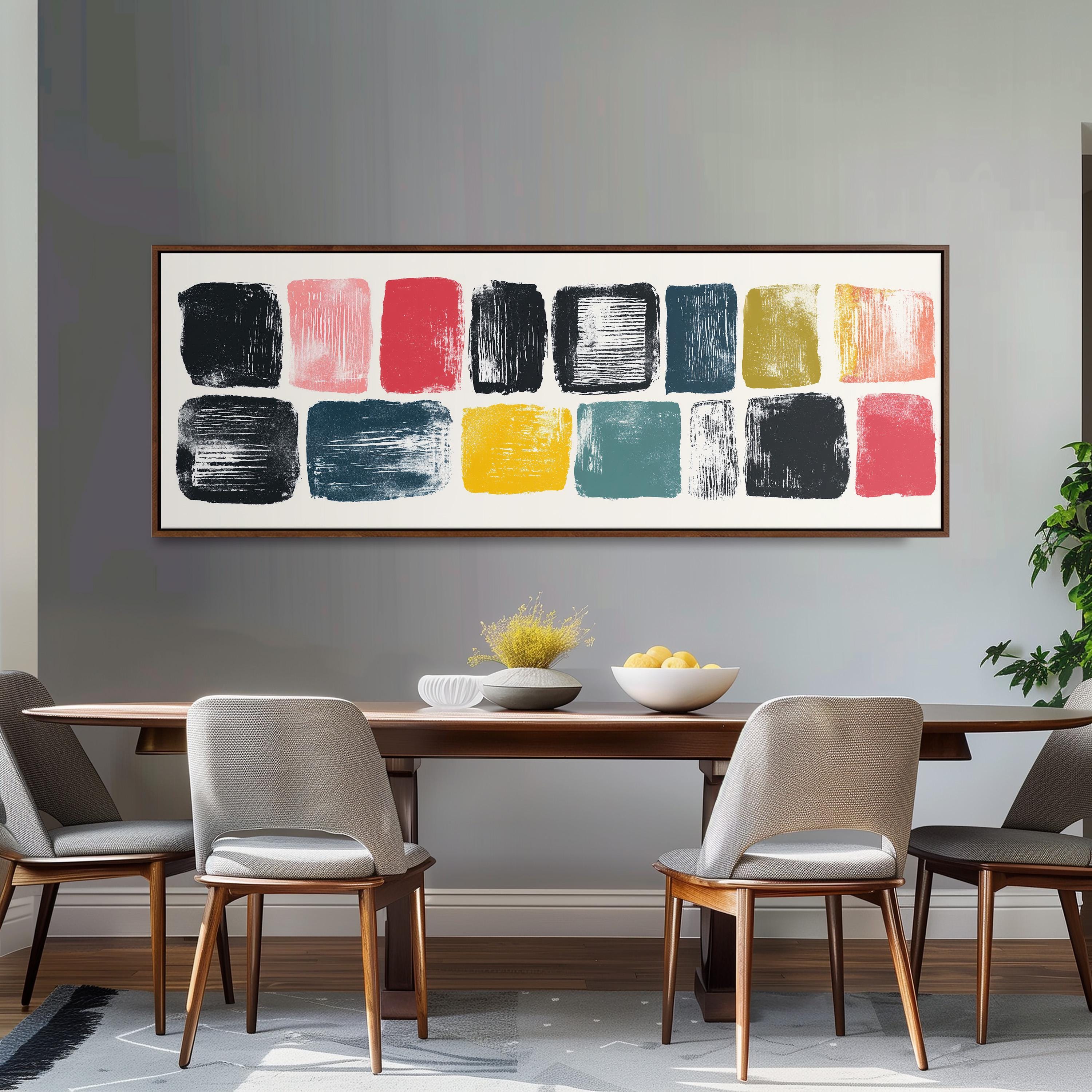a modern dining room with a wooden table and four chairs. Above the table, there is a large abstract painting with various colored squares. The table is set with a bowl of fruit and a vase of flowers, creating a welcoming and stylish atmosphere.