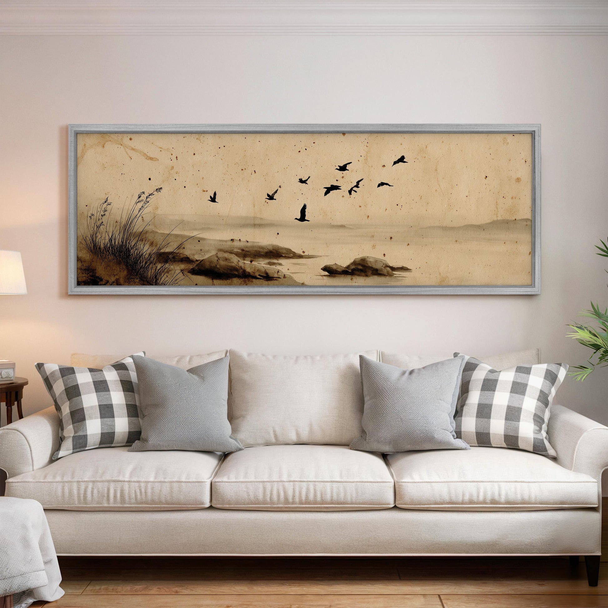 a large framed painting of a serene landscape with birds flying over a body of water, hanging on a wall above a white sofa with patterned throw pillows.