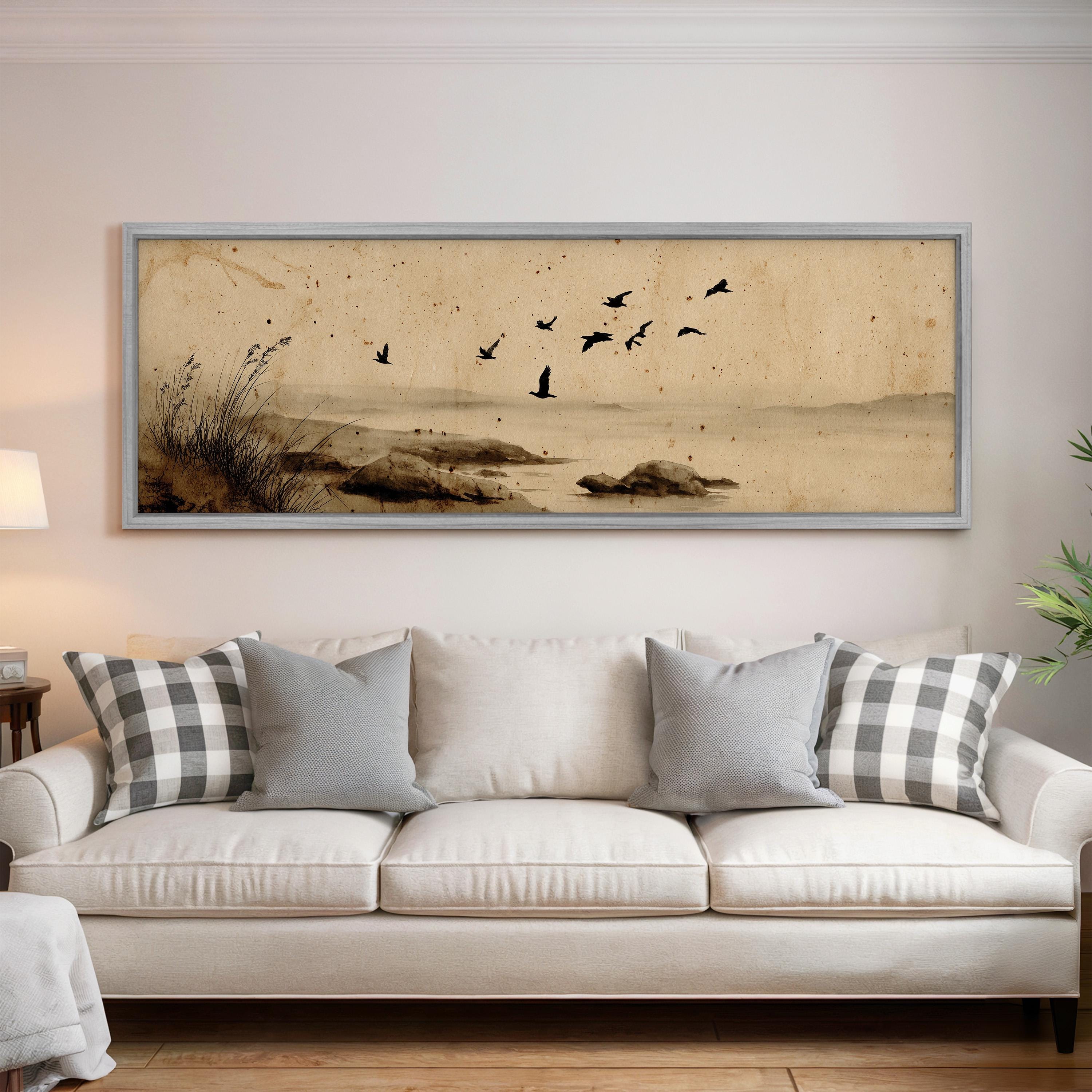 a large framed painting of a serene landscape with birds flying over a body of water, hanging on a wall above a white sofa with patterned throw pillows.