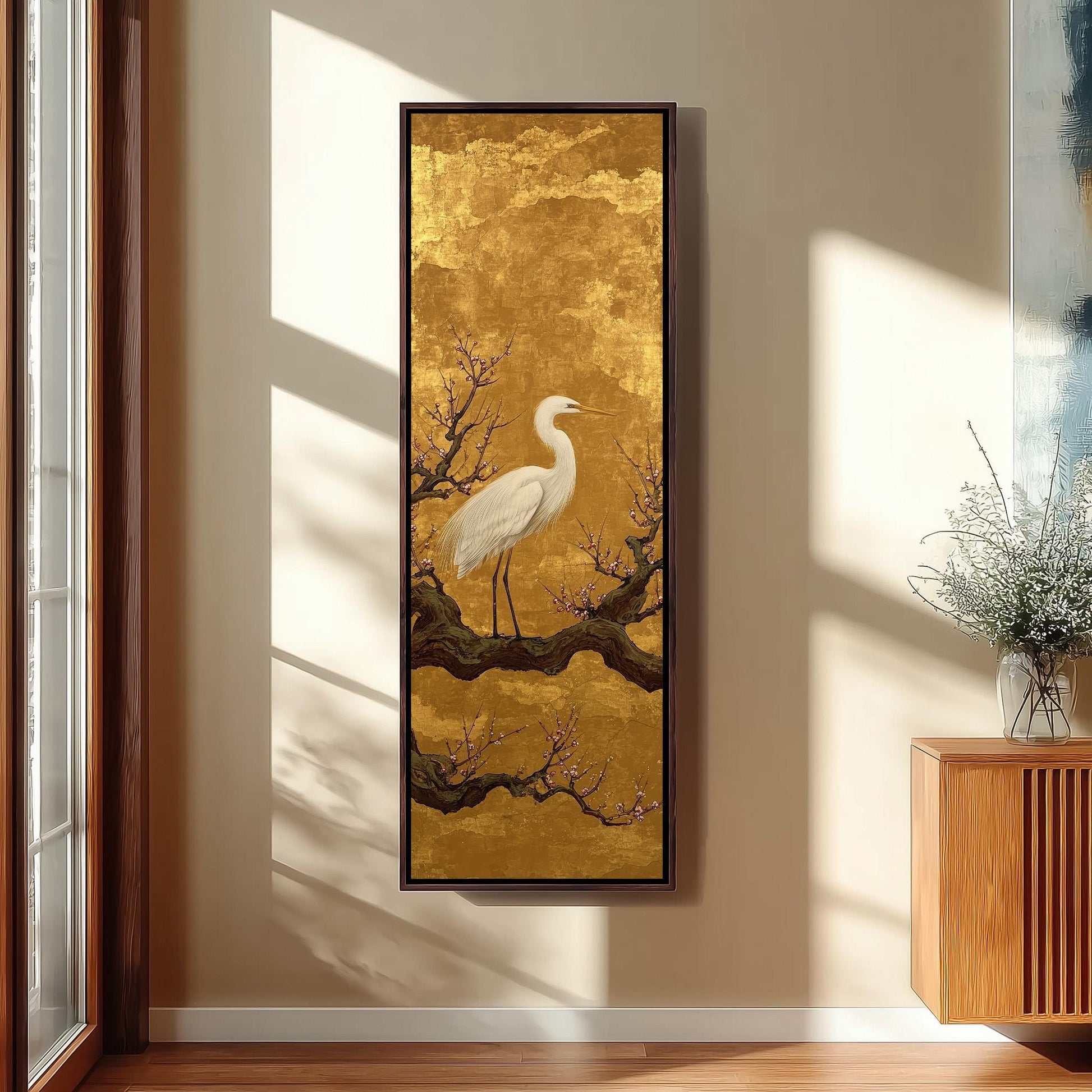 a large framed painting of a white crane standing on a branch with flowers, hanging on a wall in a room with a wooden cabinet and a vase of flowers.