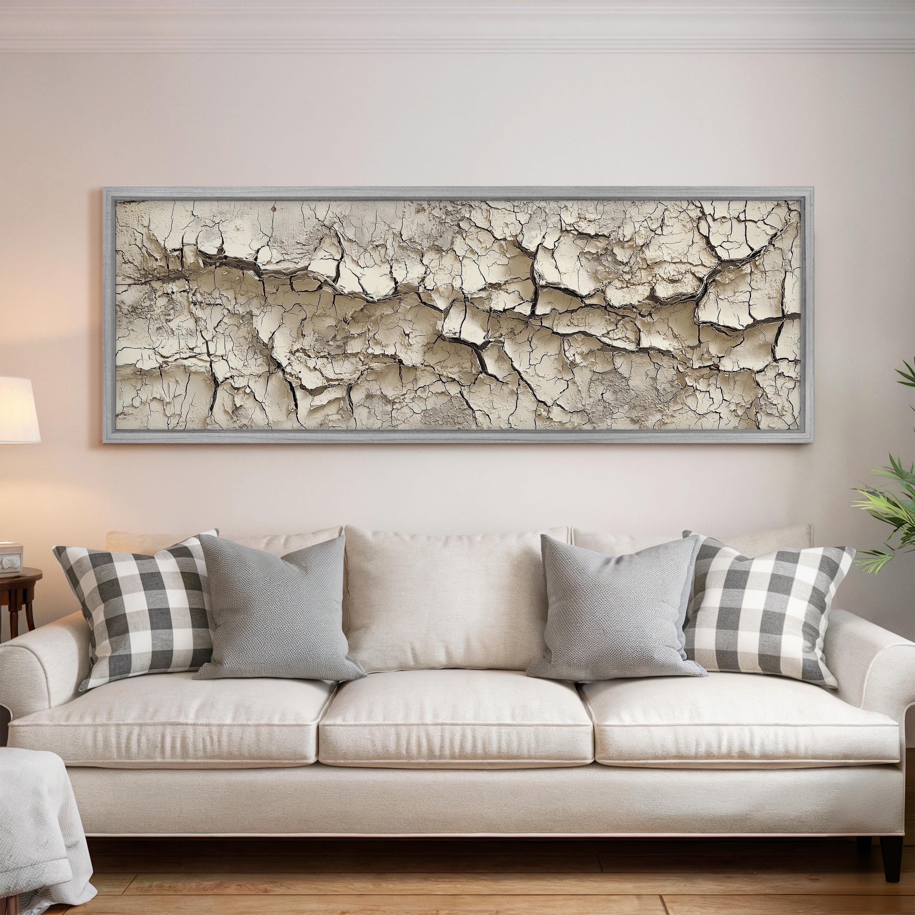 a large, white, three-seater sofa with gray and white plaid throw pillows. Above the sofa, there is a large, framed picture of cracked, pale-colored rock or stone.
