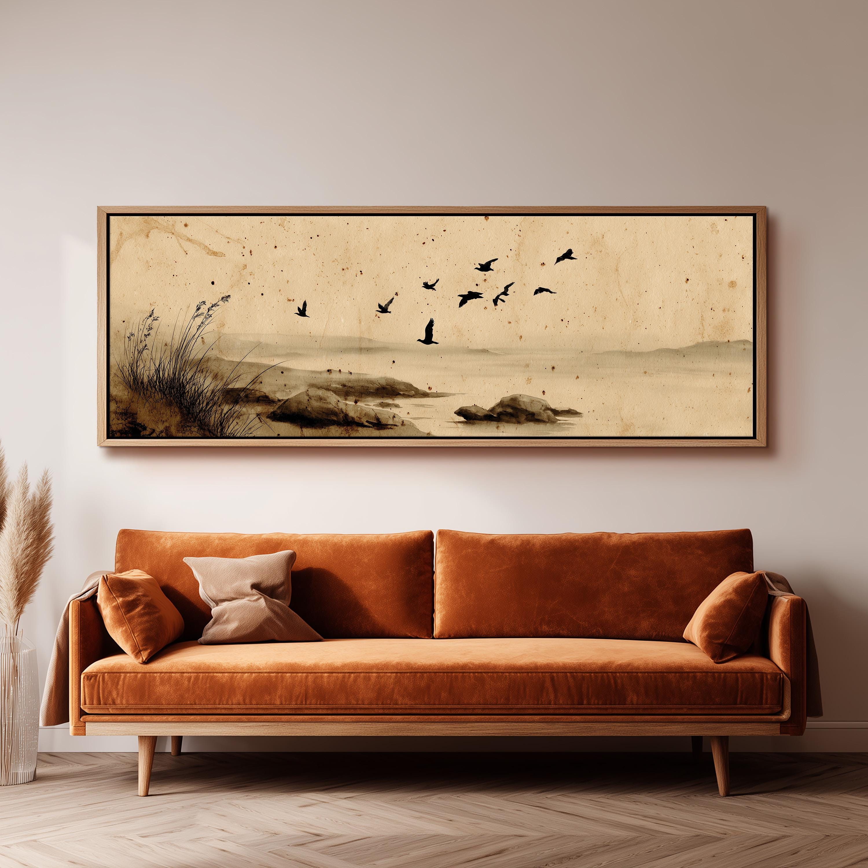 a large, framed painting of a serene landscape with birds flying over a body of water, hanging on a wall above a brown sofa.