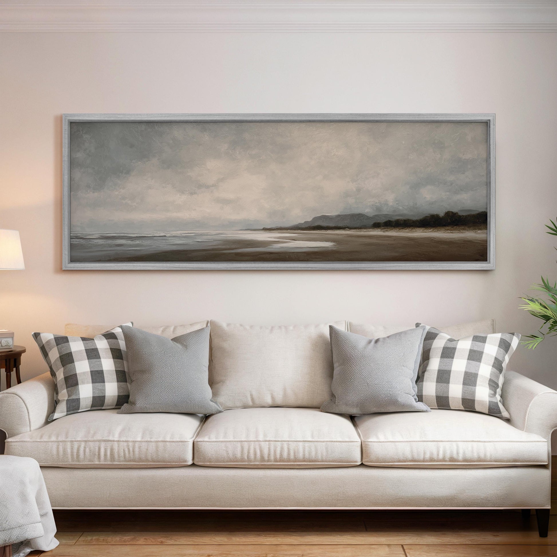 a large, framed painting of a beach landscape hanging on a wall above a white sofa with gray and white patterned throw pillows.