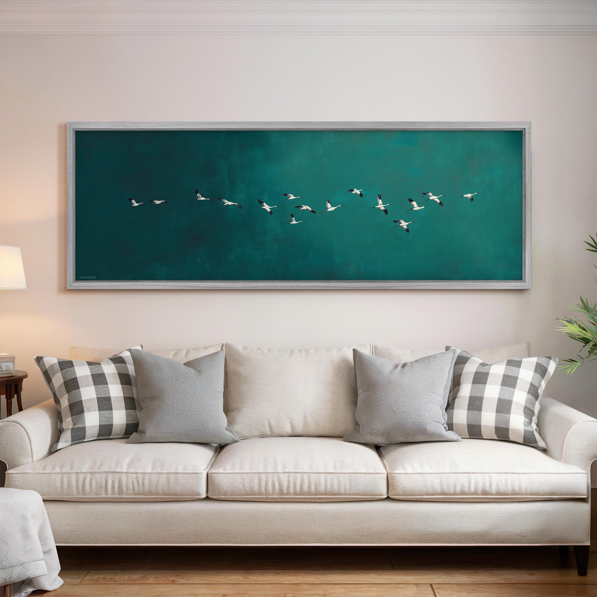 a large framed painting of a flock of birds flying over a green background, hanging on a wall above a white couch with gray and white patterned pillows.