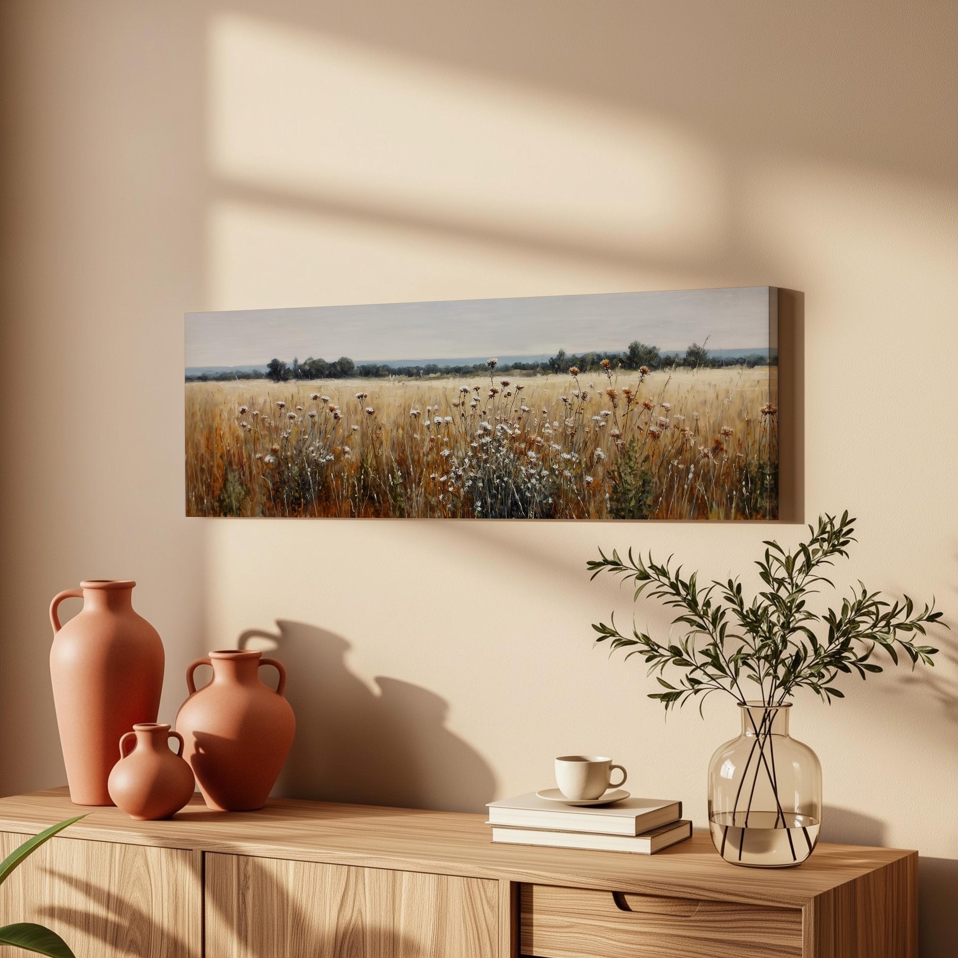 a wooden cabinet with a vase of greenery, a coffee cup, and a stack of books on top. Above the cabinet, there is a painting of a field with tall grass and wildflowers.