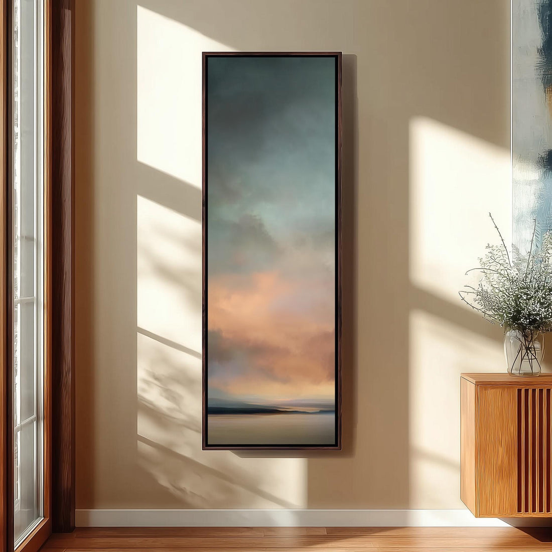 a large, rectangular painting hanging on a wall, depicting a serene landscape with a sunset or sunrise. The painting is framed and placed in a room with a wooden cabinet and a vase of flowers on a nearby table.