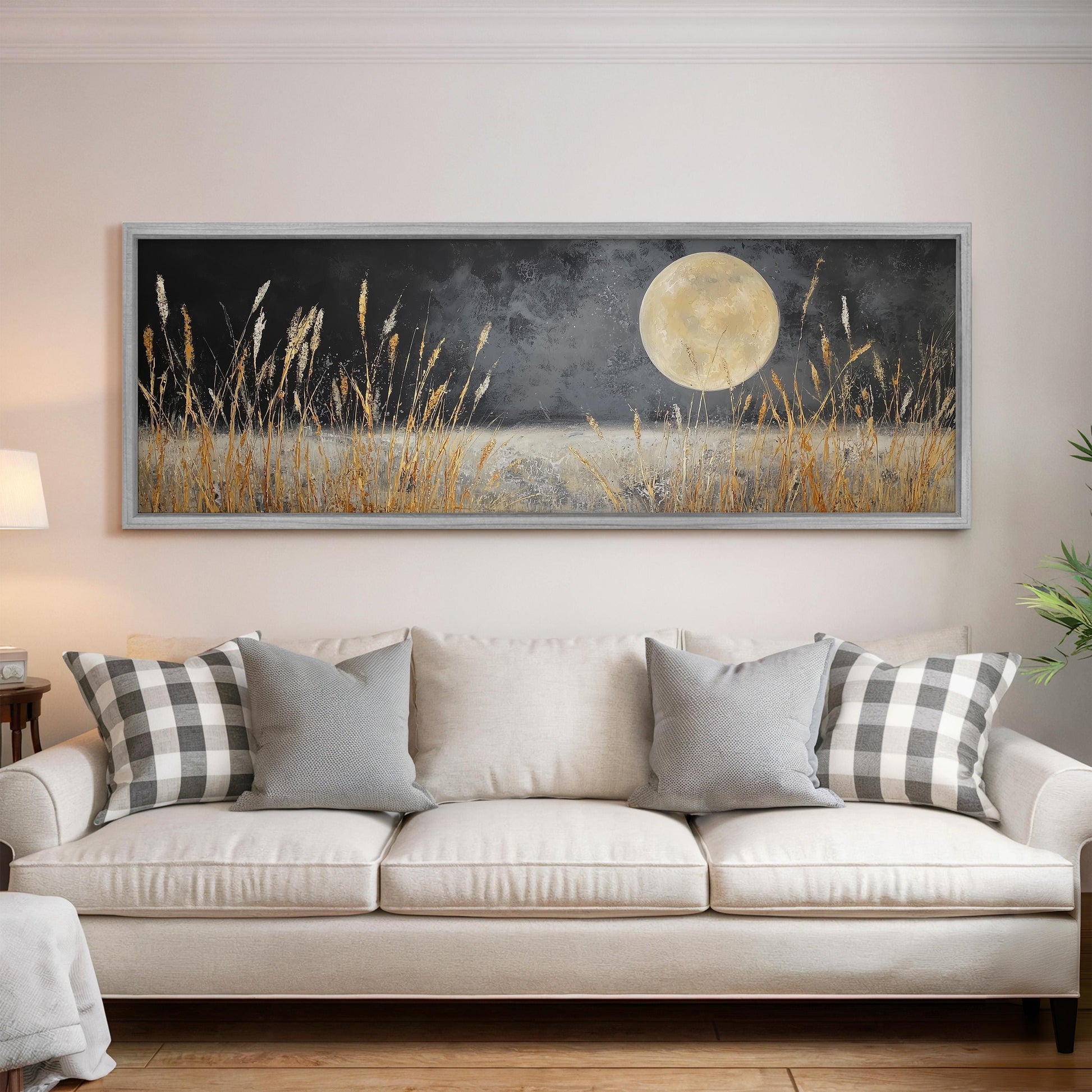 a large, framed painting of a night scene with a full moon and tall grasses in the foreground, hanging on a wall above a white couch with gray and white patterned pillows.