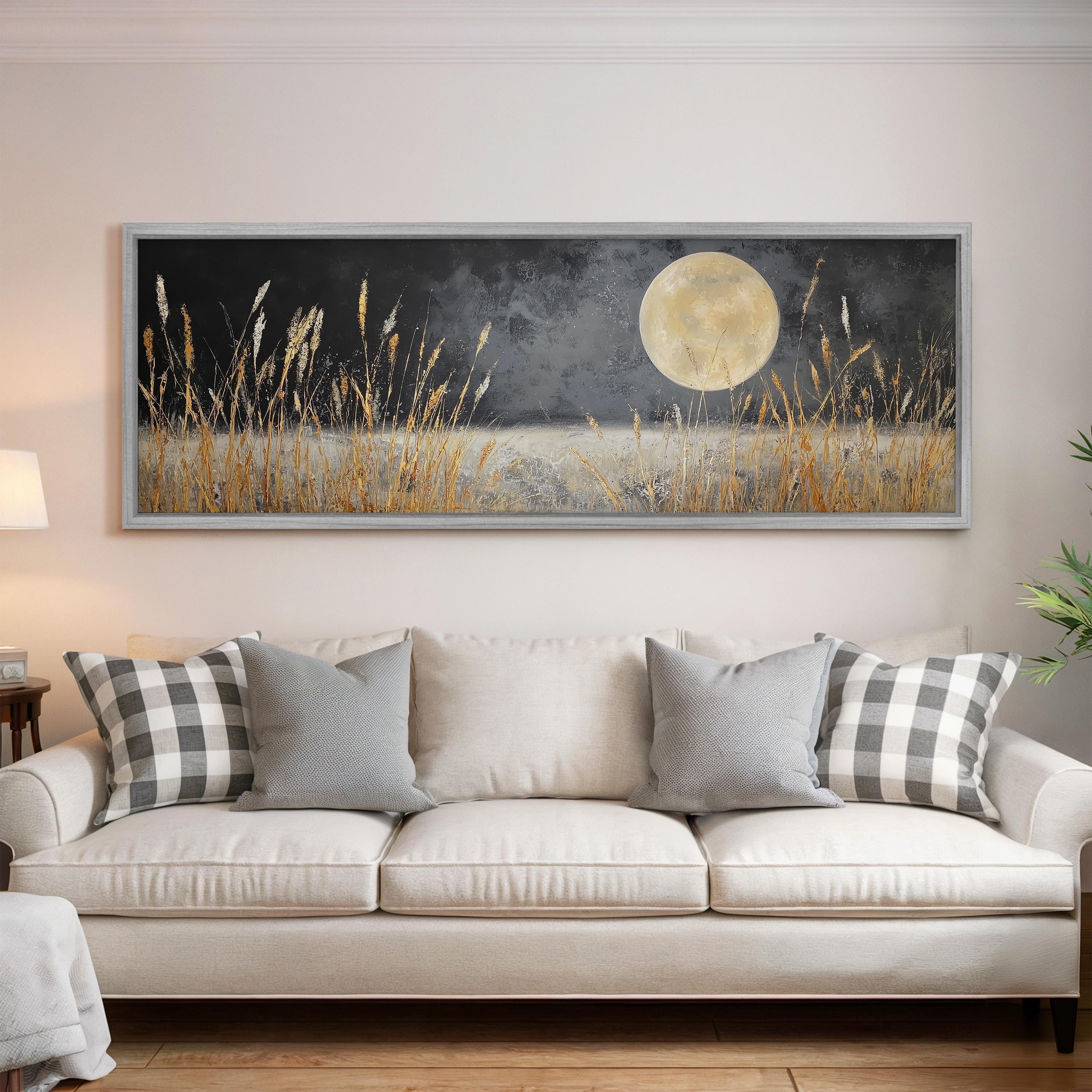 a large, framed painting of a night scene with a full moon and tall grasses in the foreground, hanging on a wall above a white couch with gray and white patterned pillows.