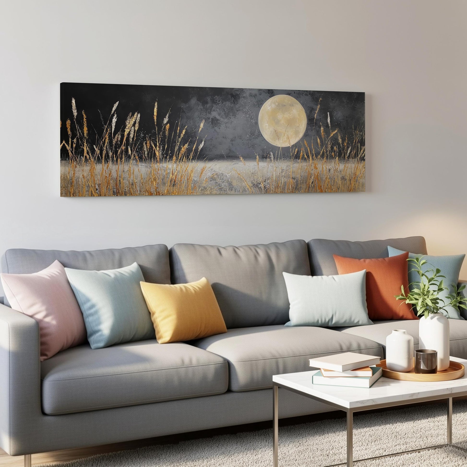 a living room with a gray sofa adorned with colorful throw pillows, a coffee table, and a painting of a field with tall grass and a full moon hanging on the wall above the sofa.