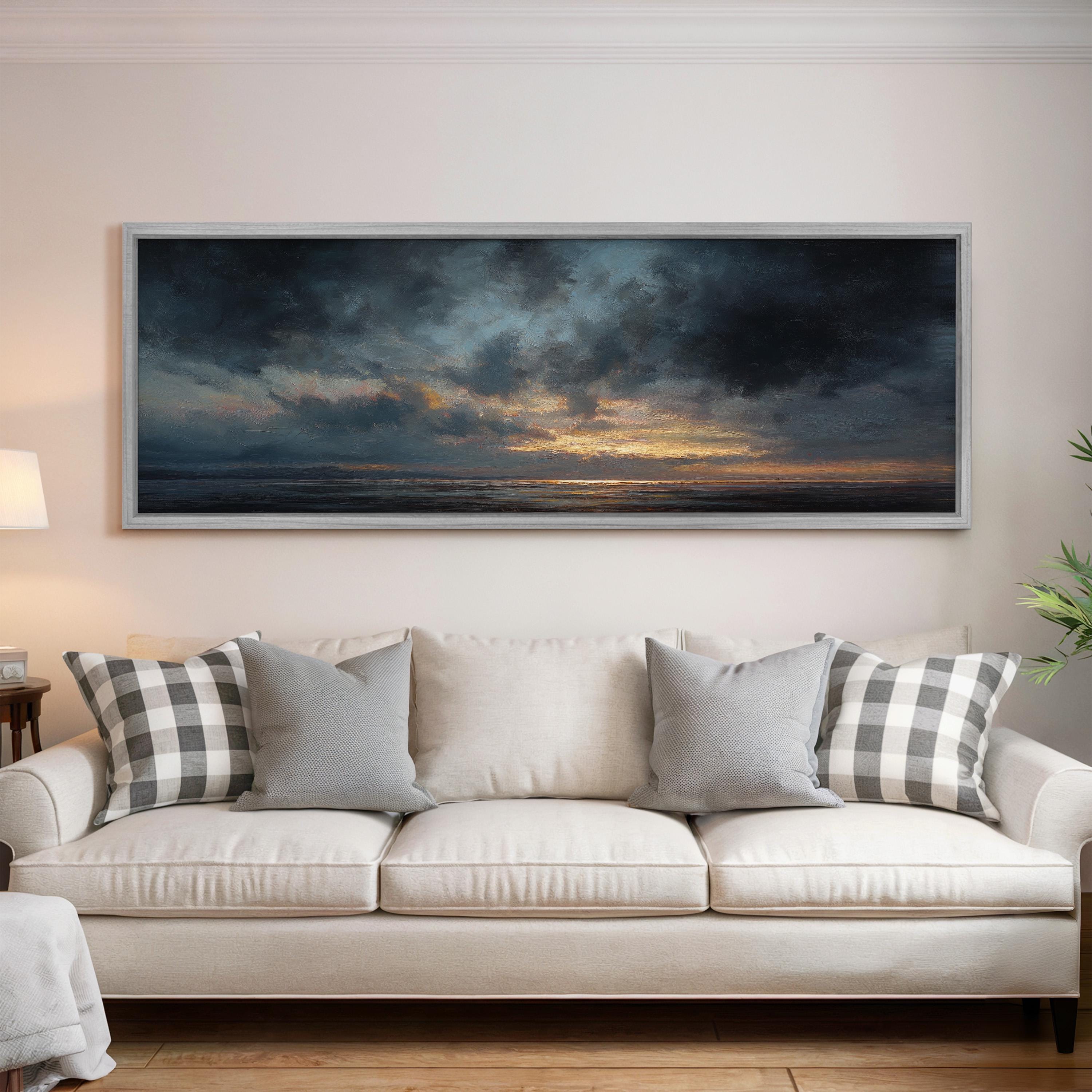 a large, framed painting of a dramatic sky with clouds and a sunset, hanging on a wall above a white sofa with patterned throw pillows.