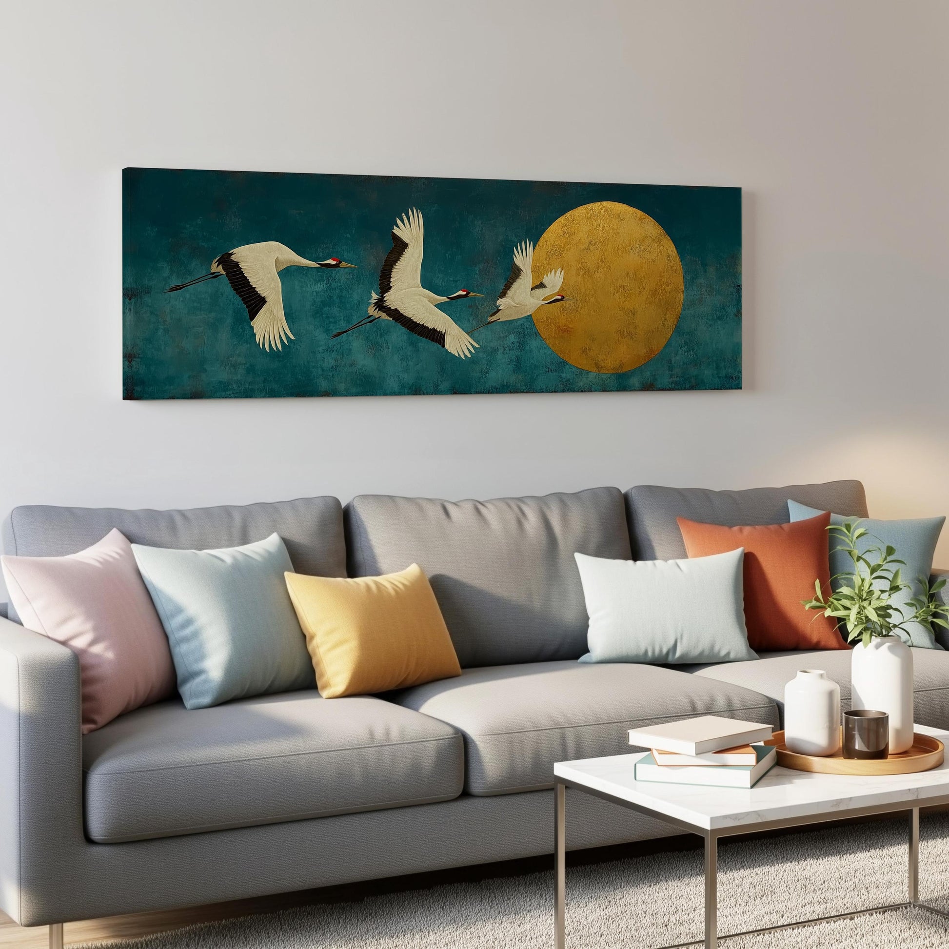 a large painting of three white birds in flight against a blue background, with a large yellow circle in the center. The painting is hanging on a wall above a gray couch with colorful throw pillows. In the foreground, there is a white coffee table with a stack of books and a small plant.