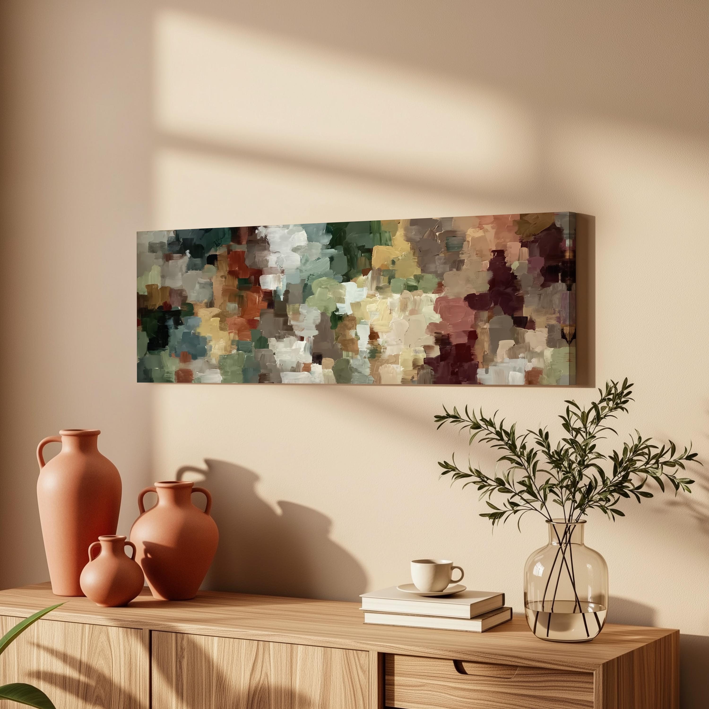 a wooden cabinet with a painting hanging on the wall above it, along with three orange vases and a glass vase with greenery. The cabinet also has a stack of books and a coffee cup on top.