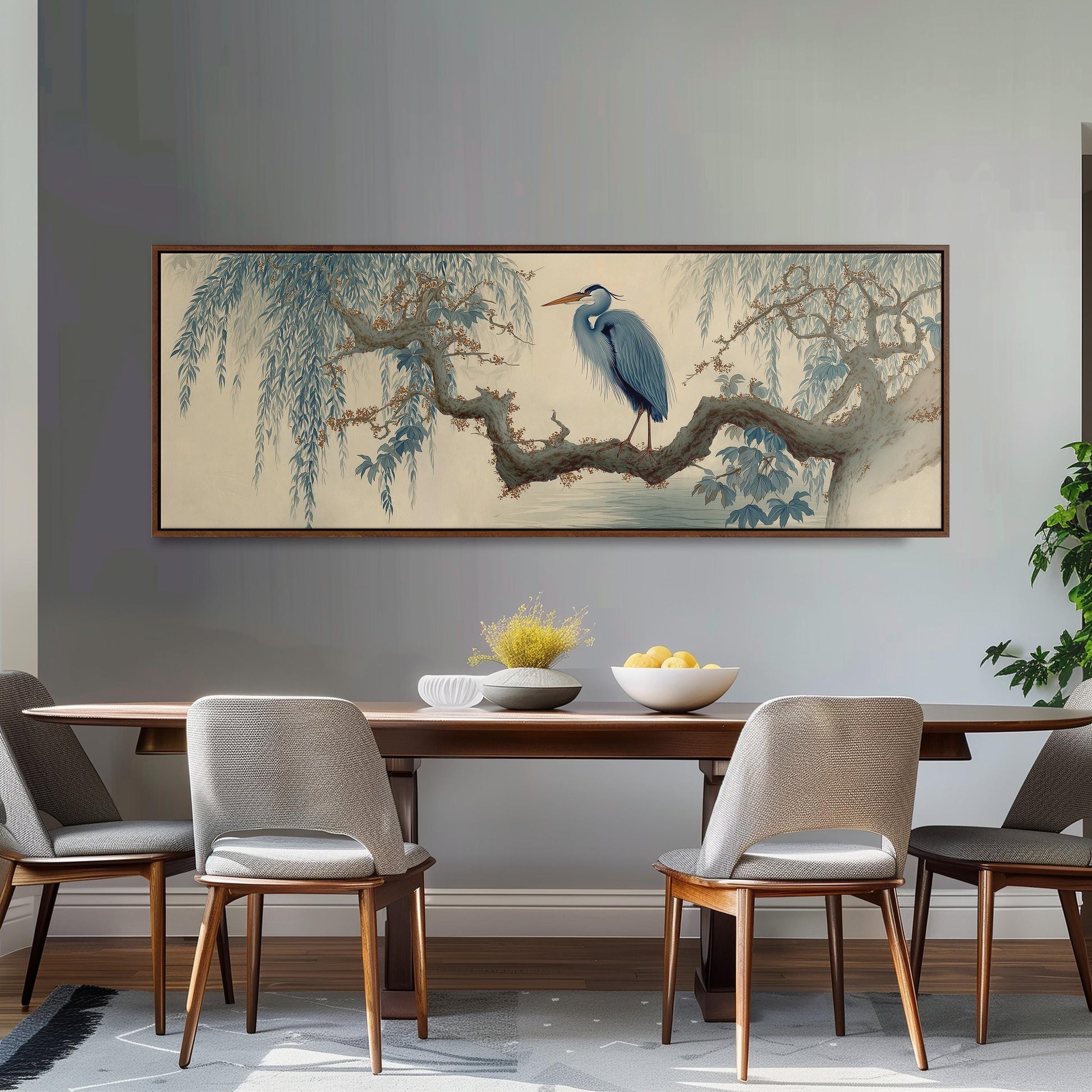 a dining room with a large painting of a blue heron perched on a branch above a wooden table. The table is set with a bowl of fruit and a vase of flowers. The room has gray walls and a gray carpet, creating a calm and elegant atmosphere.