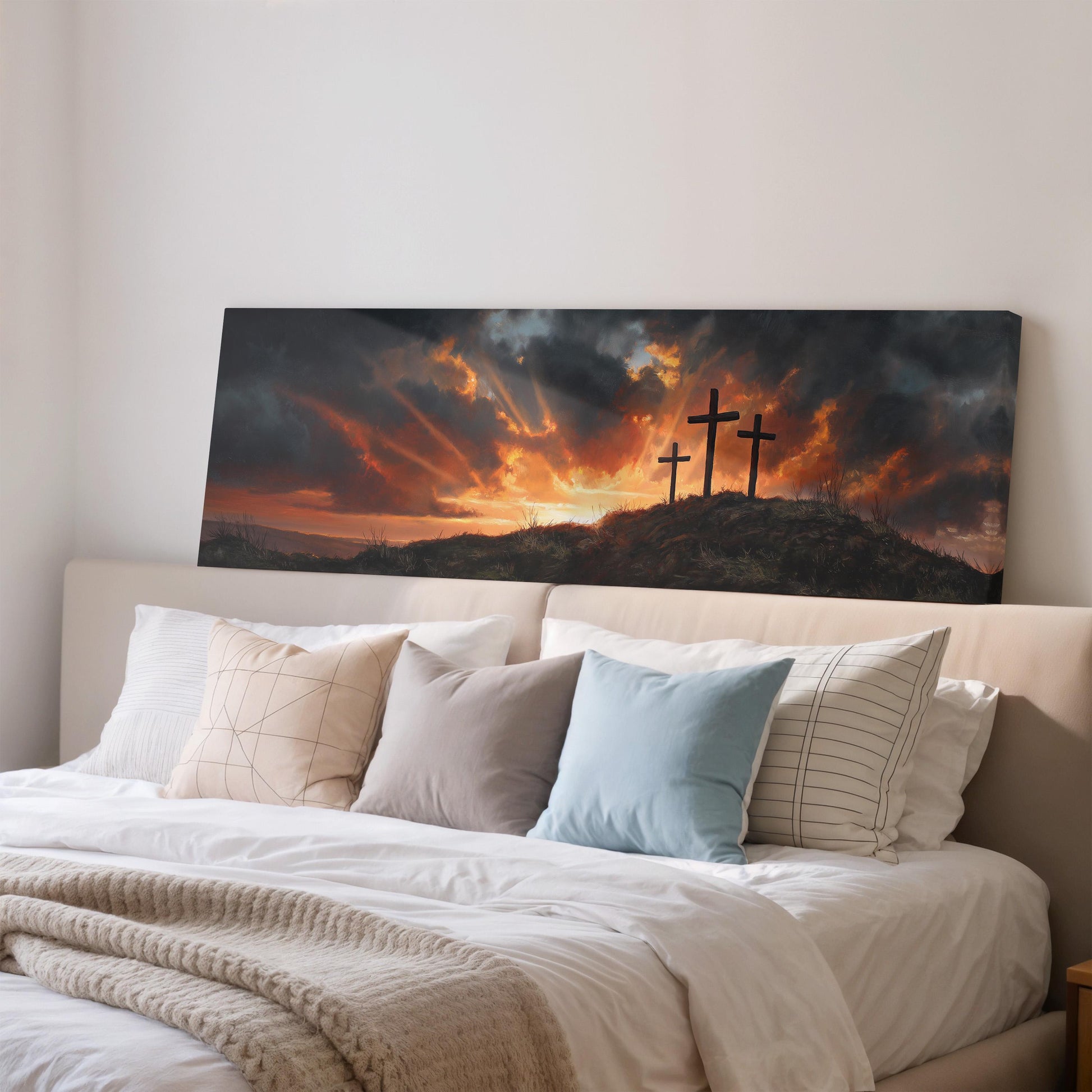 a serene bedroom scene with a large bed adorned with white linens and a blue pillow. Above the bed, a painting of a dramatic landscape with three crosses silhouetted against a fiery sunset sky is displayed.