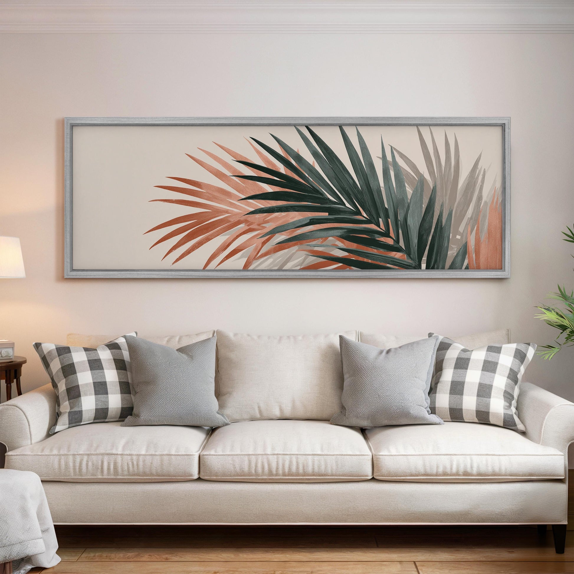 a living room with a white sofa adorned with gray and white plaid throw pillows. Above the sofa, there is a large framed picture of a palm leaf with a mix of orange and green colors. The room has a clean and minimalist aesthetic, with a white wall and a wooden floor.