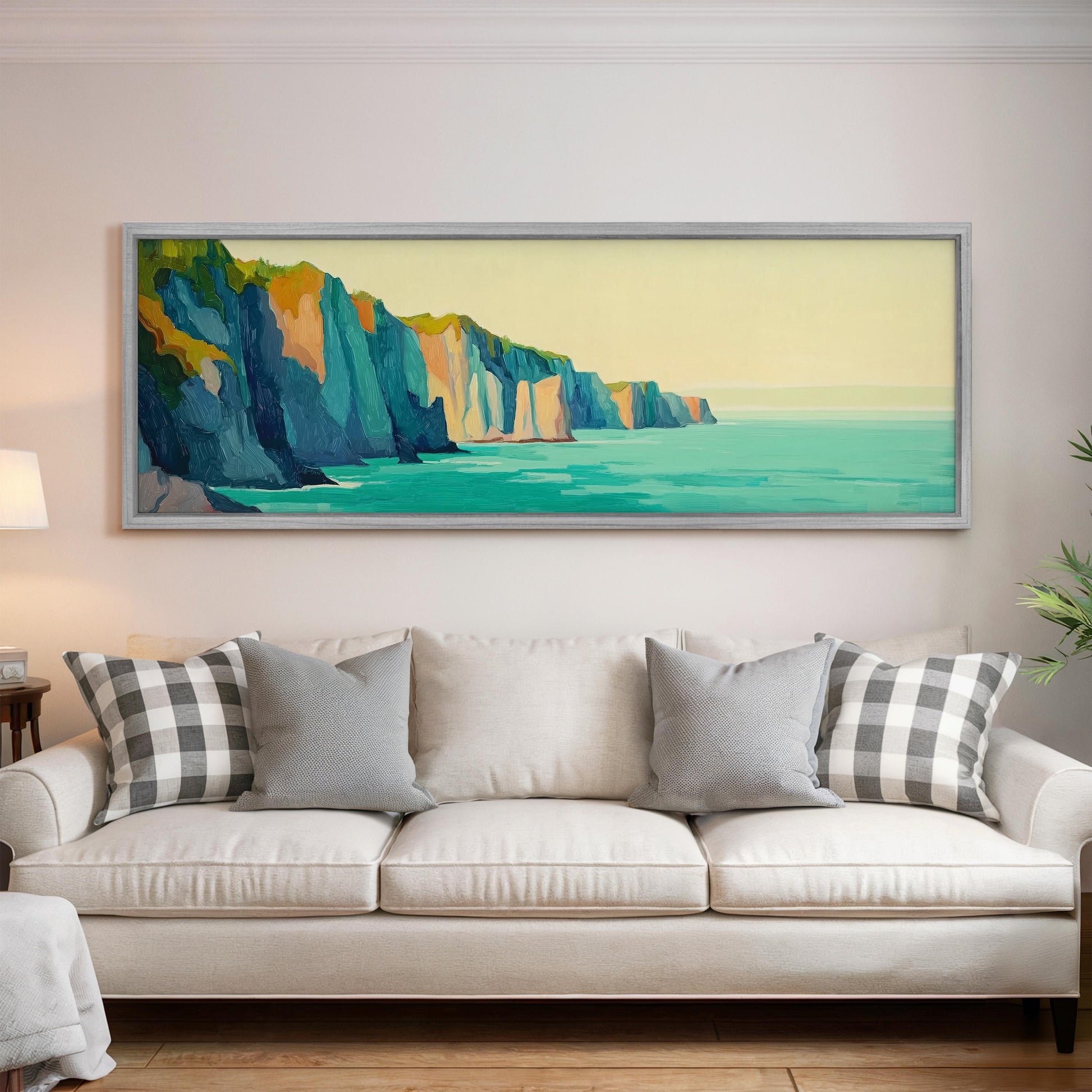 a large, framed painting of a cliff overlooking the ocean, hanging on a wall above a white couch with gray and white patterned pillows.