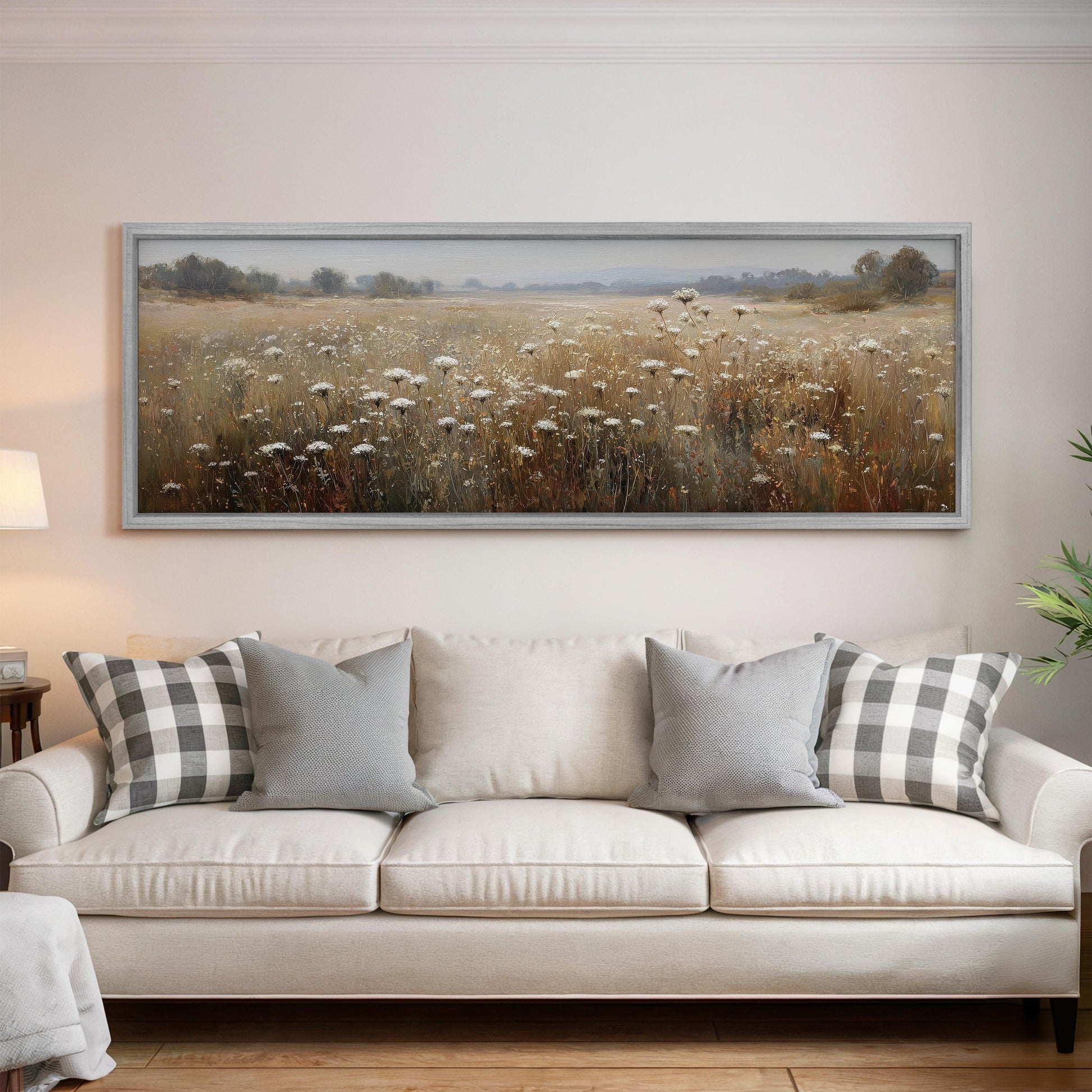 a large, framed painting of a field with tall grass and wildflowers hanging on a wall above a white sofa with gray and white patterned pillows.
