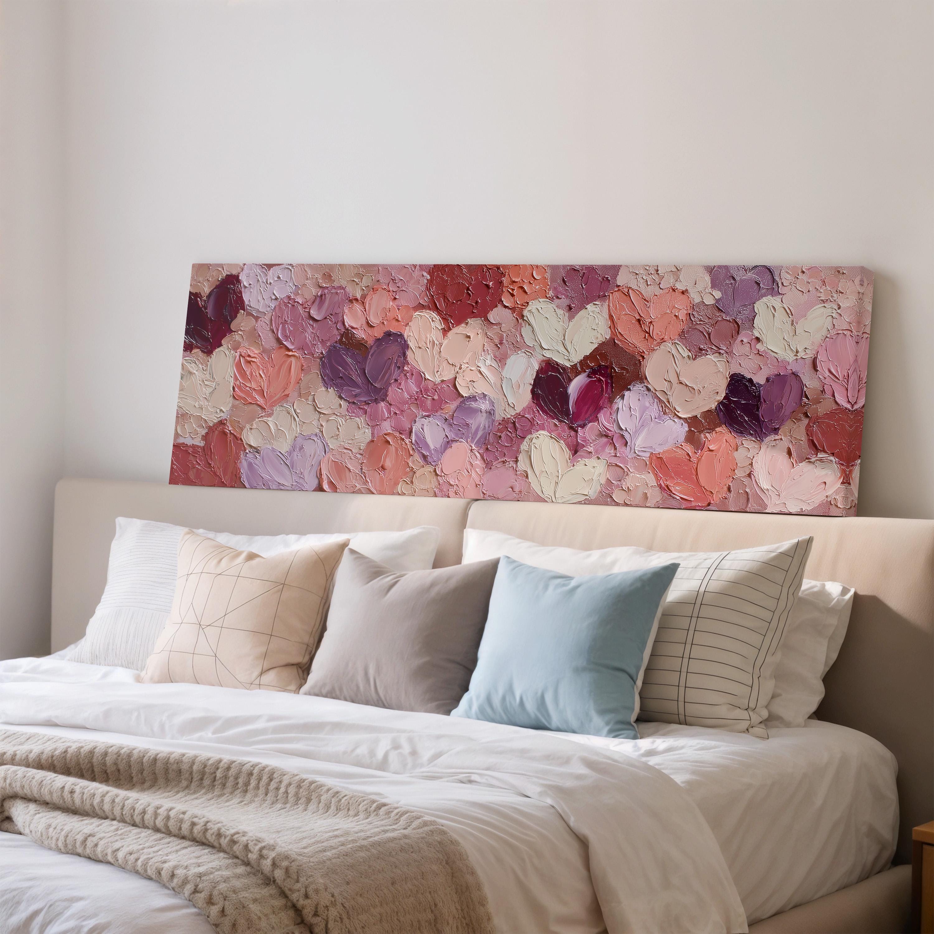 a large bed with a white comforter and a variety of colorful pillows. Above the bed, there is a large painting of hearts in different shades of pink and purple.