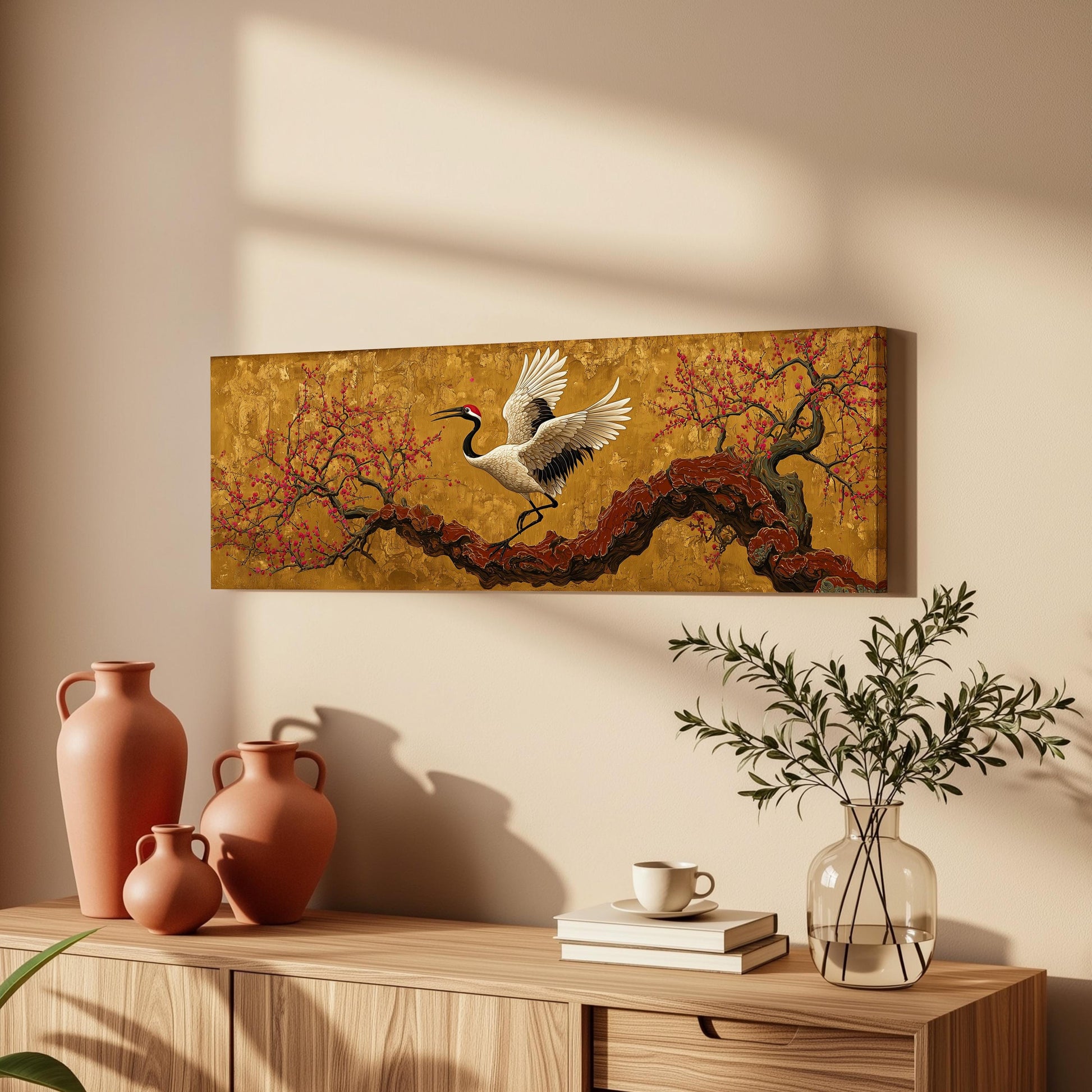 a wooden cabinet with a vase, a coffee cup, and a stack of books on top. Above the cabinet, there is a framed painting depicting a crane in flight over a tree with red leaves.